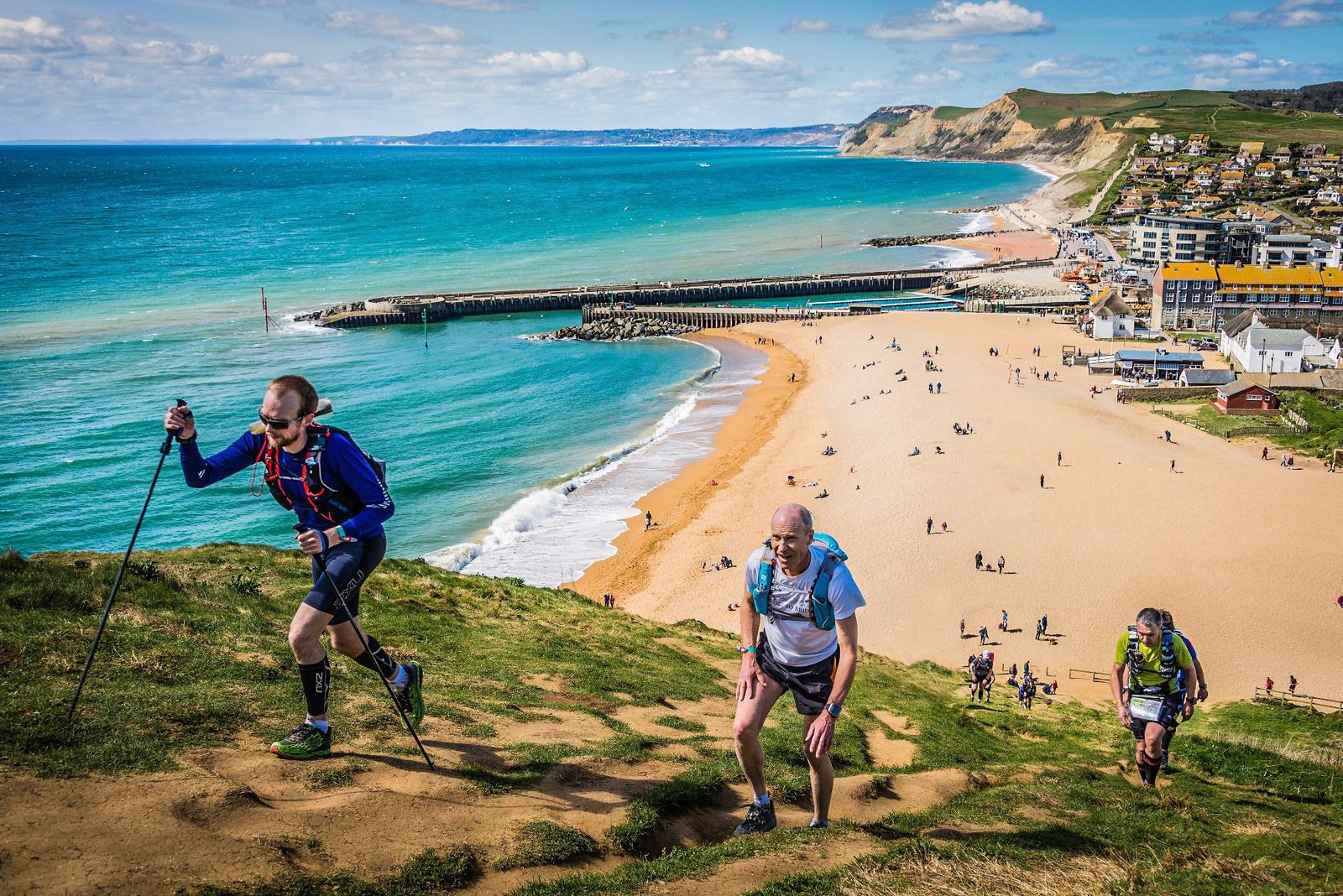 Three people are hiking on a coastal trail with a scenic view of a beach, ocean, and distant cliffs. One person uses trekking poles. The sky is partly cloudy, and there are people on the beach and near buildings in the background.