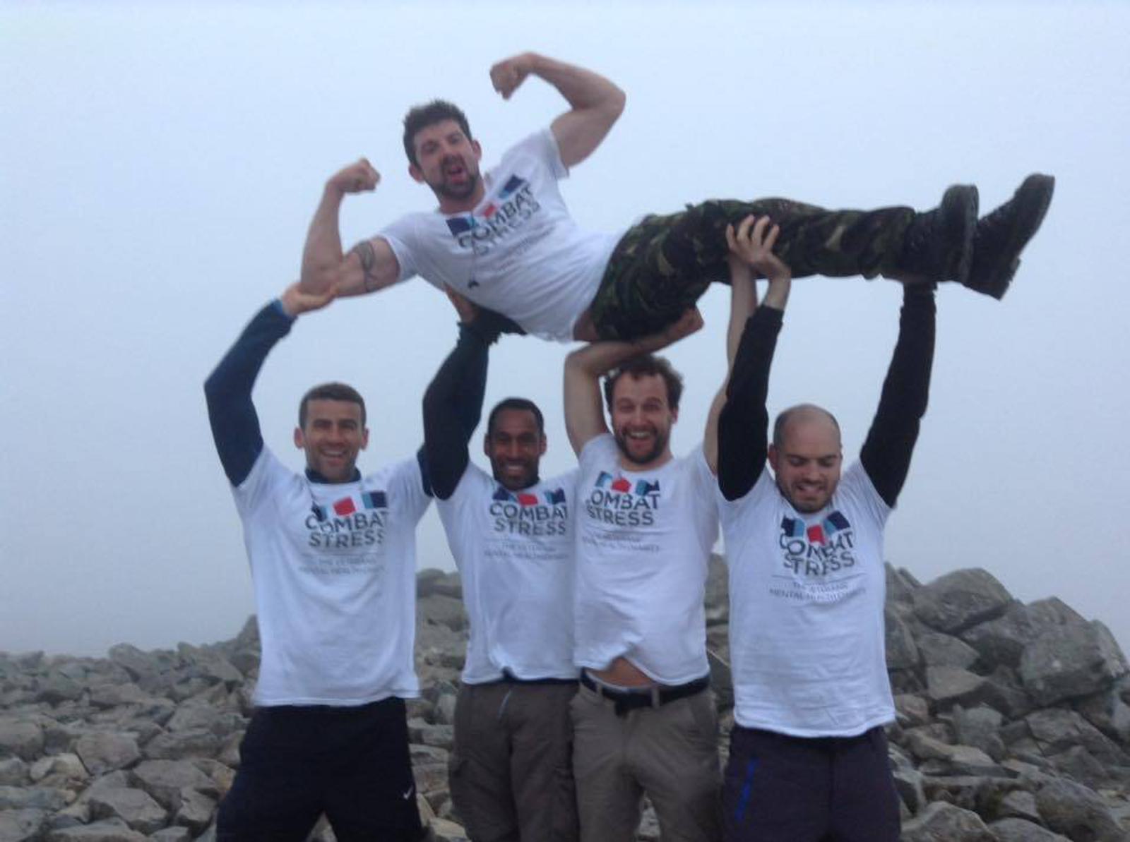 Five men wearing "Combat Stress" T-shirts are posing on a rocky landscape. Four are standing, with two lifting another man horizontally above them. They appear joyful and energetic, with one man flexing his muscles. The sky is foggy.