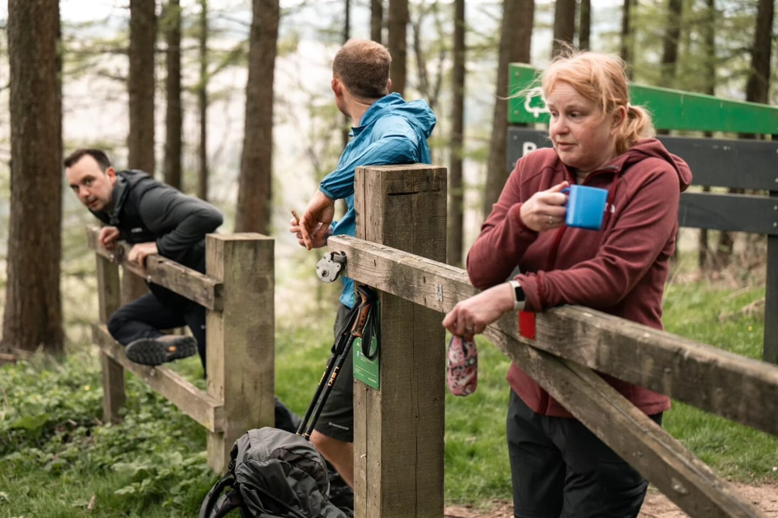 Three people pause by a wooden fence in a forest; one woman in a red hoodie drinks from a blue mug while two men rest, with one climbing over the fence and the other looking back.
