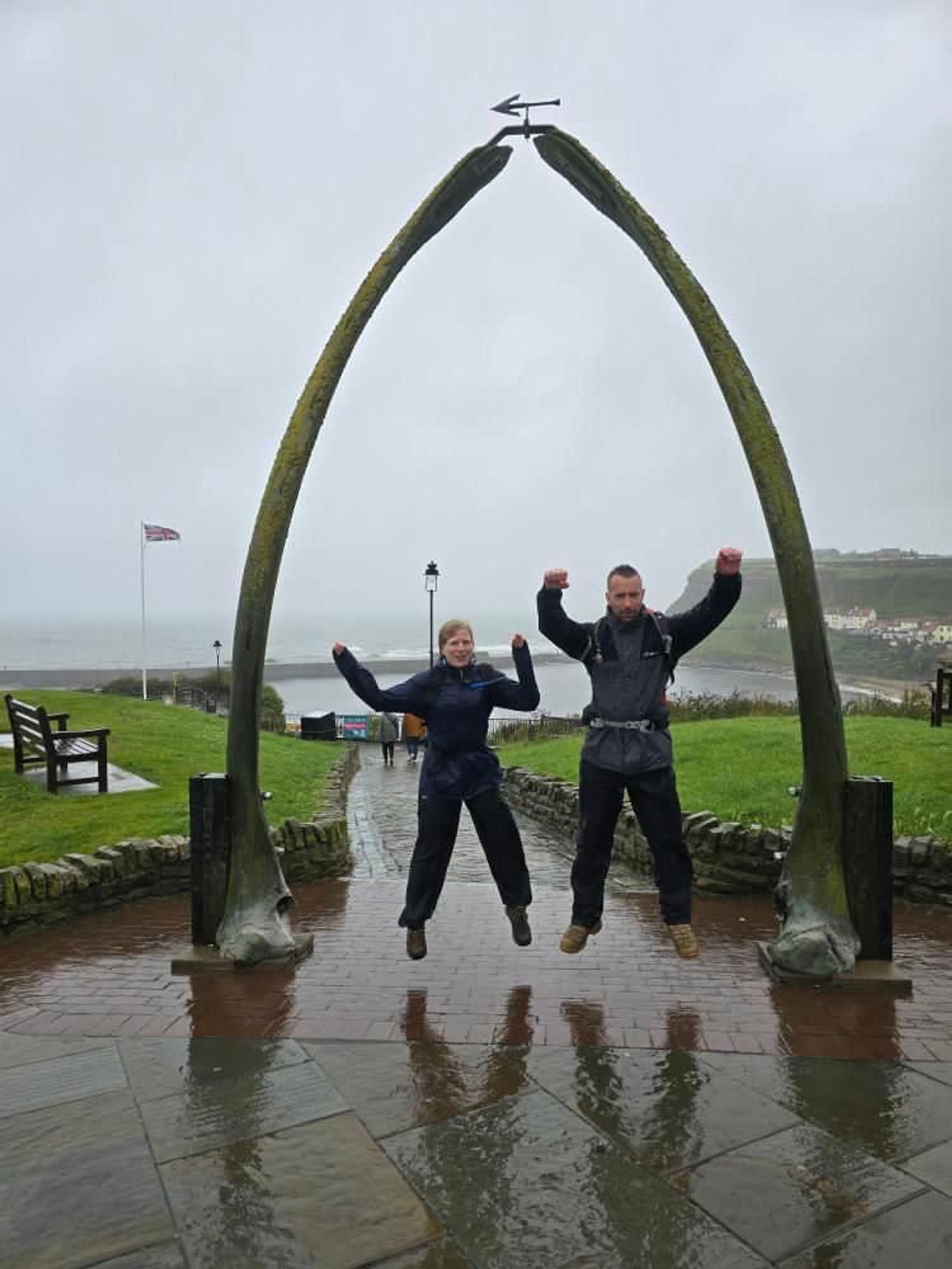Two people jumping under a large, arch-shaped sculpture on a rainy day. The wet ground reflects their images. In the background, there's a grassy area, a few benches, a path, and a distant view of the sea with a cloudy sky.