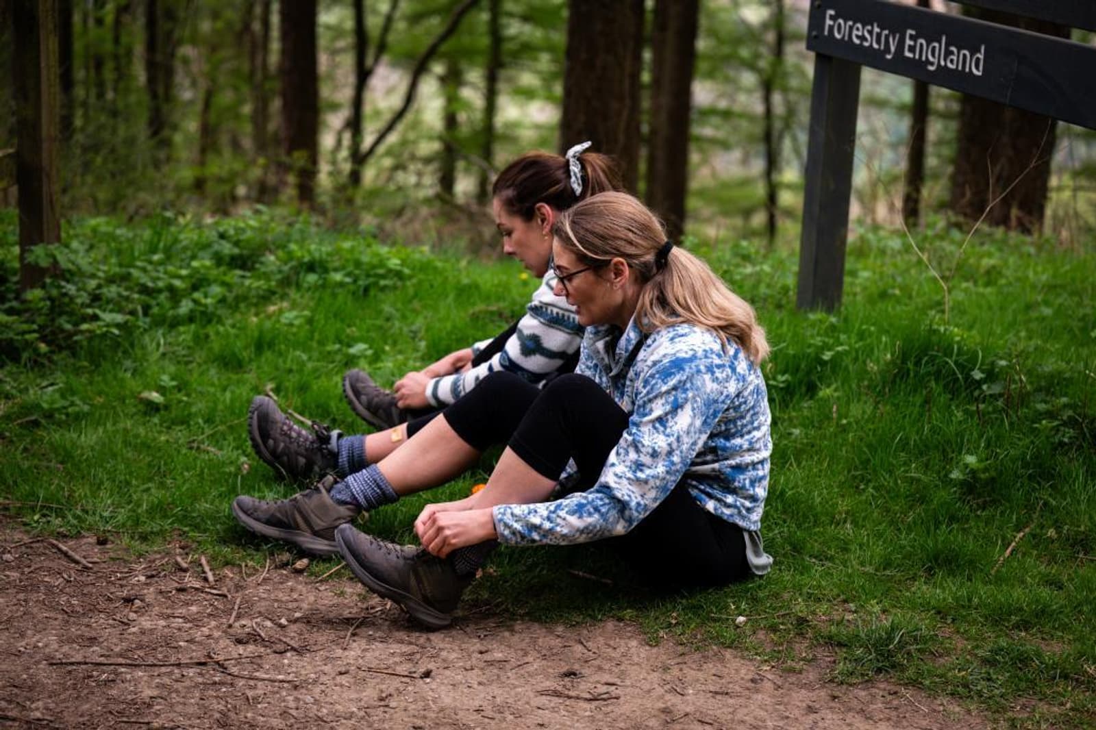 Two women sit on the grass in a forest, adjusting their hiking boots. There is a "Forestry England" sign nearby, and the surrounding area is green and wooded.