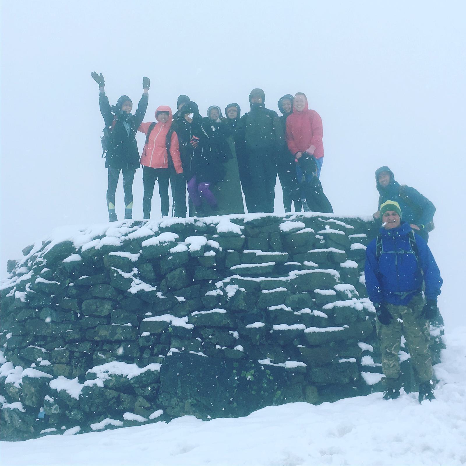 A group of ten people in winter clothing stand on a snow-covered stone structure. Some are raising their arms in celebration. The background is foggy, giving a sense of cold and adventure.