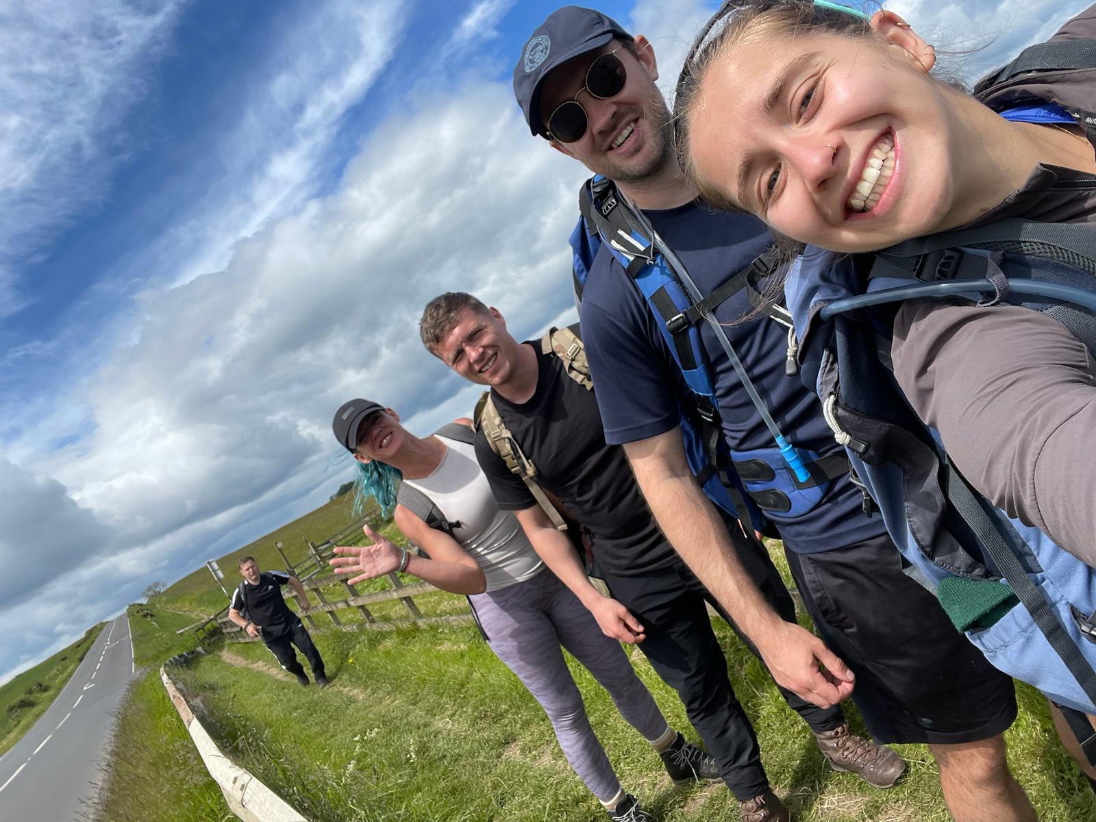 Four people are hiking along a grassy path beside a road, smiling and posing for a selfie. They are dressed in outdoor gear, with backpacks. The sky is partly cloudy, and another person walks behind them.