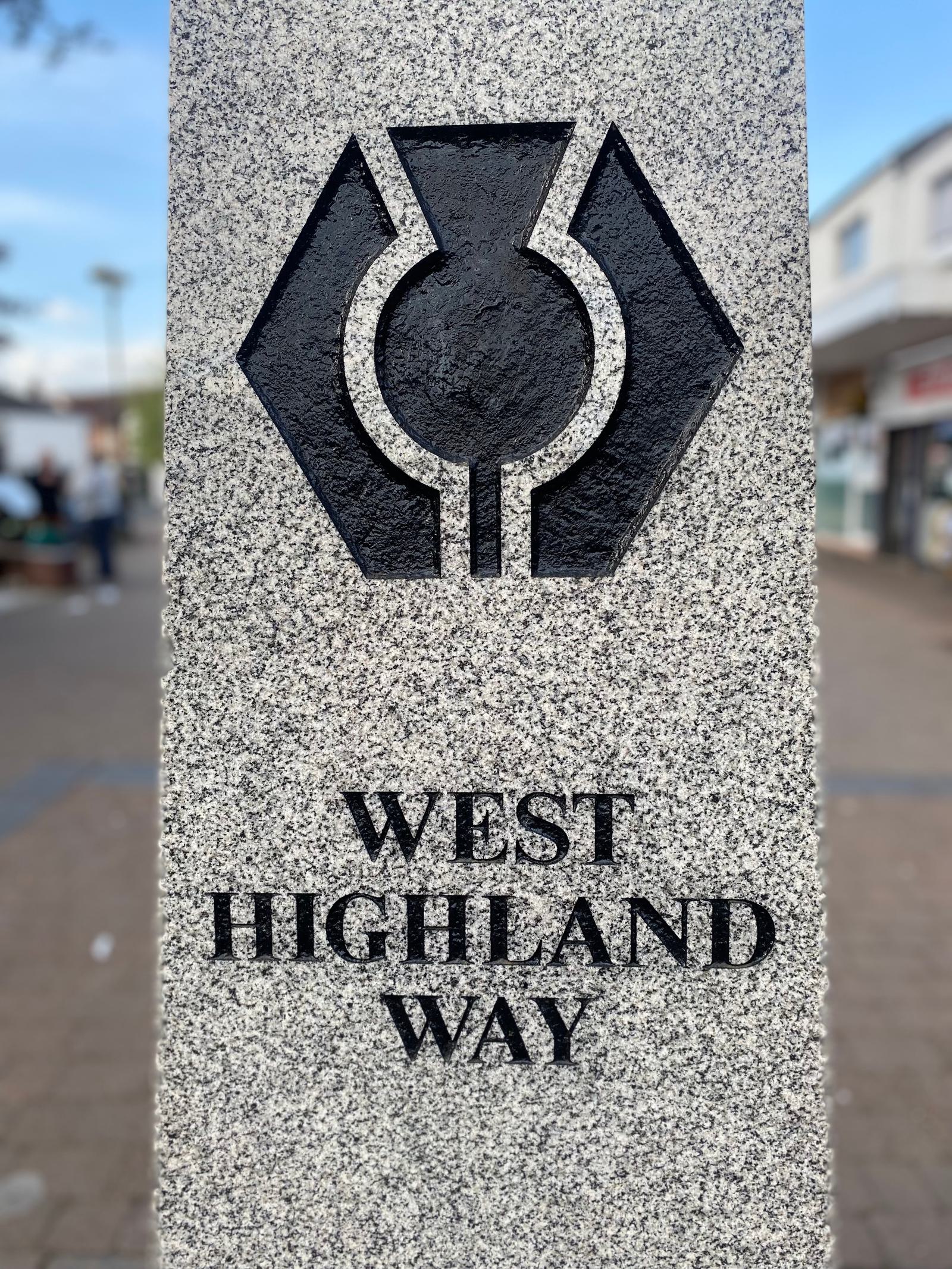 Stone marker for the West Highland Way featuring a black geometric logo. The text is etched below the logo. Background shows an out-of-focus street scene with buildings and people.