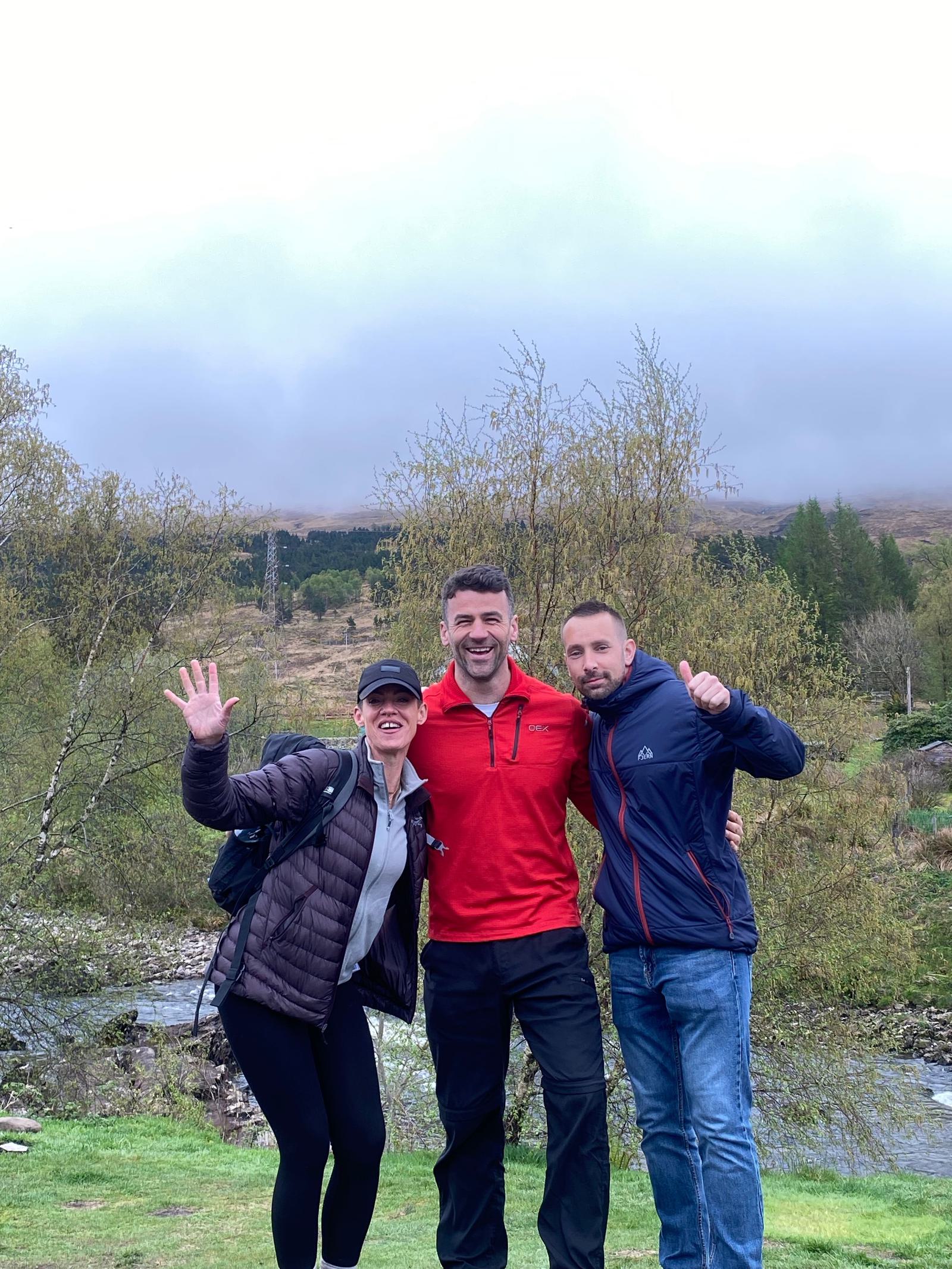 Three people stand outdoors near a small stream, smiling and waving. They are dressed in jackets and casual attire. The background features trees and a cloudy sky, suggesting a cool day.
