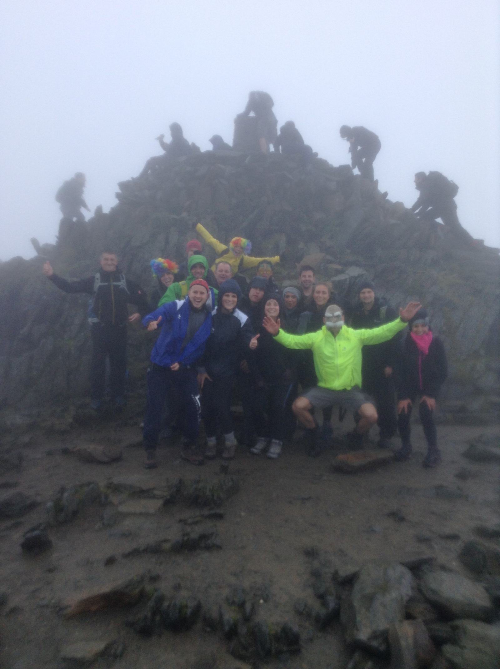 A group of people posing on a rocky, foggy mountaintop. Some are wearing colorful clothing and hats. The atmosphere is misty, with silhouettes of more people climbing in the background.