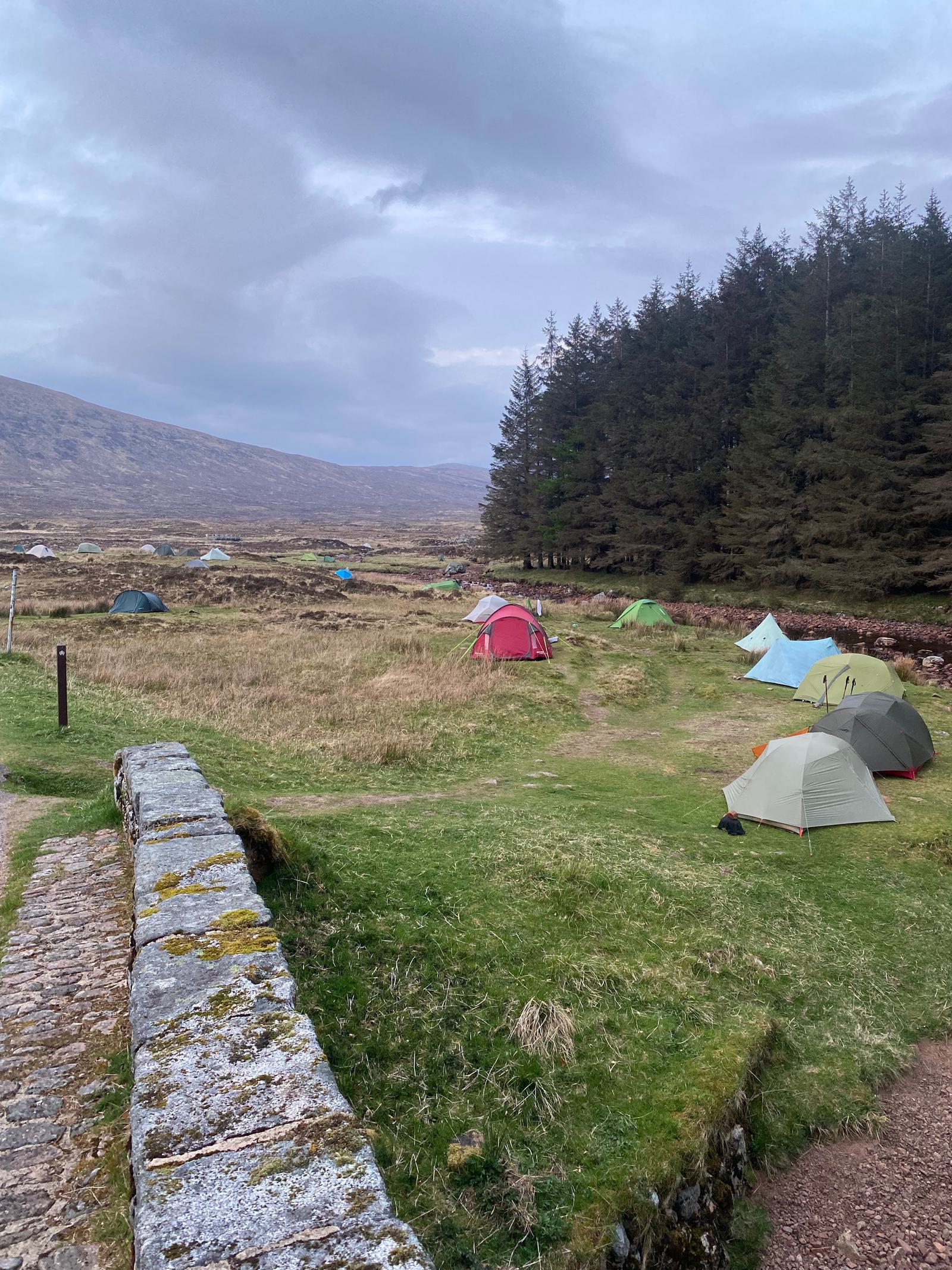 A scenic campsite with several tents scattered on a grassy field. A stone path leads through the area, bordered by a dense forest on the right and rolling hills in the background under a cloudy sky.