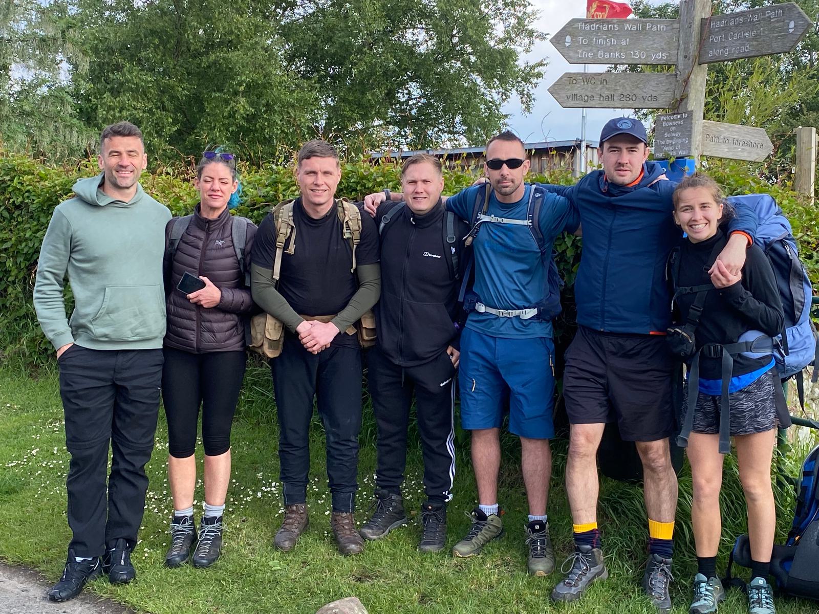 A group of seven people in outdoor clothing are standing together on a grassy area. They appear to be hikers, with backpacks and walking gear. There's a wooden signpost in the background and green foliage around them. They look cheerful and relaxed.