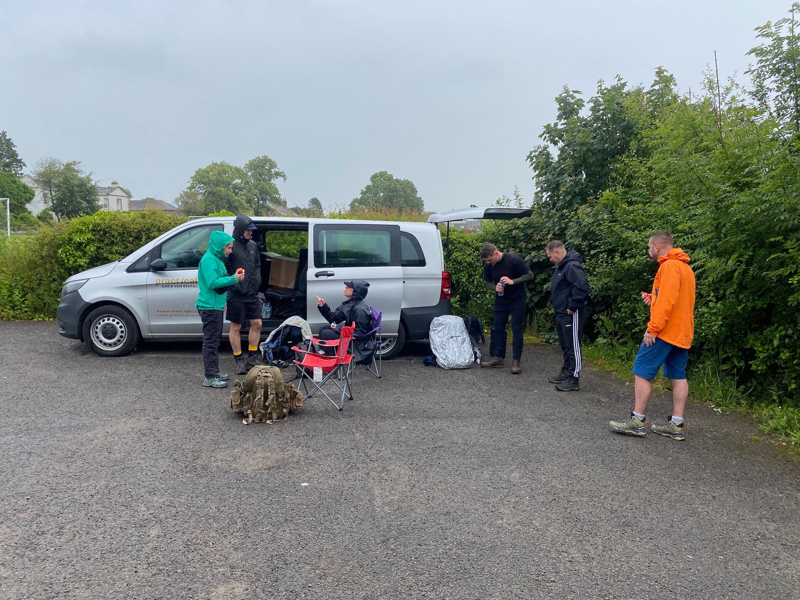 A group of people in casual outdoor clothing gather around a parked van on a cloudy day. They are standing near folding chairs and backpacks, preparing for an outdoor activity. Green trees and a building are visible in the background.