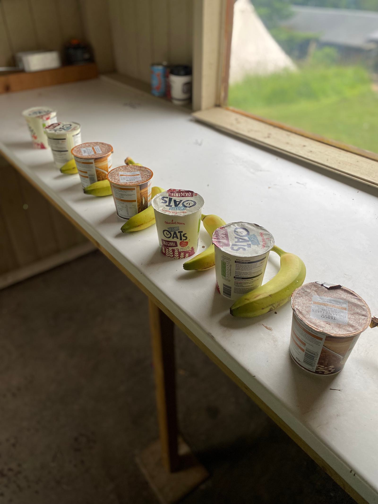 Seven yogurt containers and three bananas are lined up on a white countertop near a window, with a view of greenery outside. The scene suggests a simple breakfast setup.