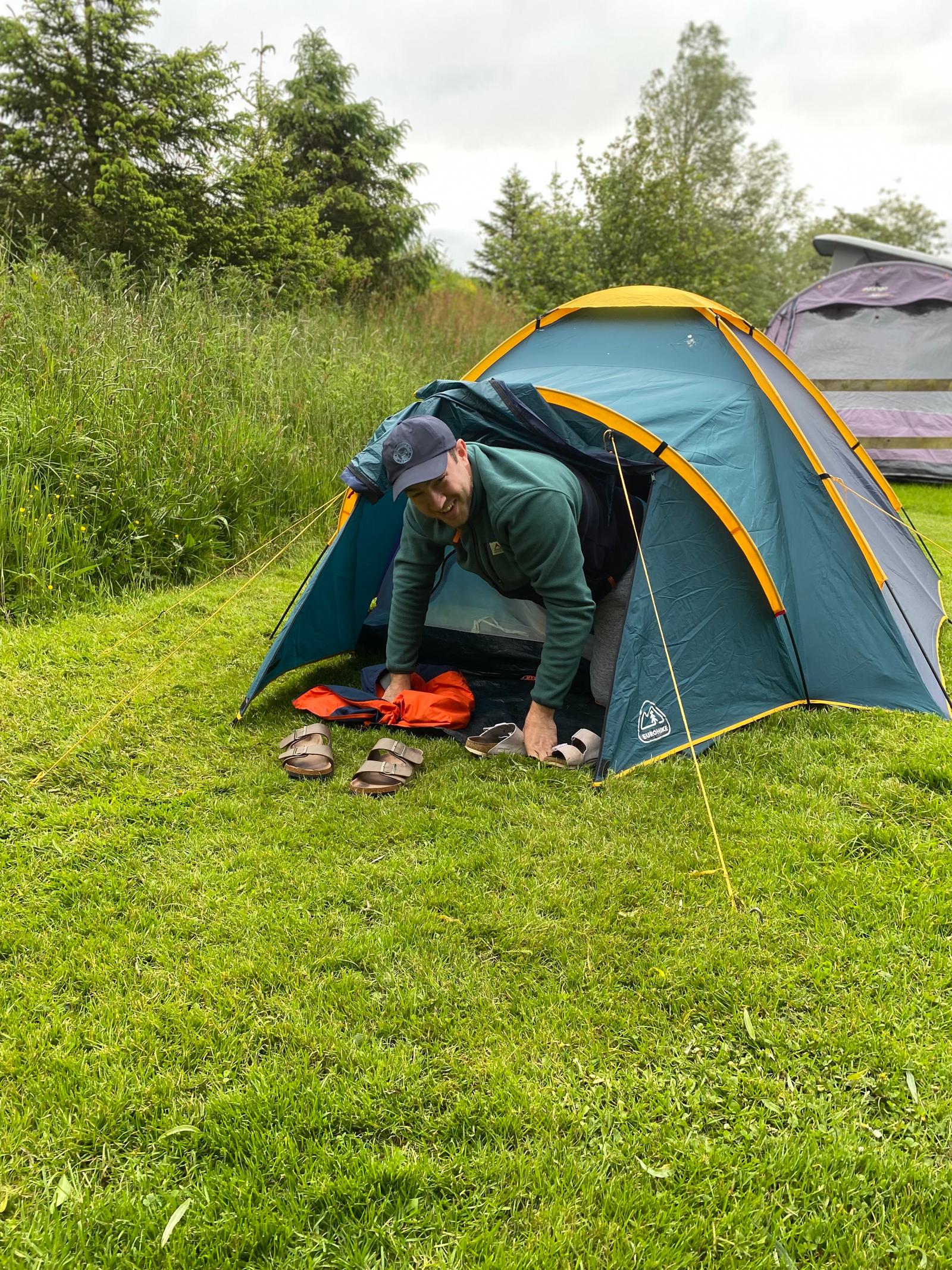 A person in a green jacket and cap is kneeling at the entrance of a small green tent pitched on grass. They are organizing belongings, including shoes and a red item, outside the tent. Other tents and trees are visible in the background.