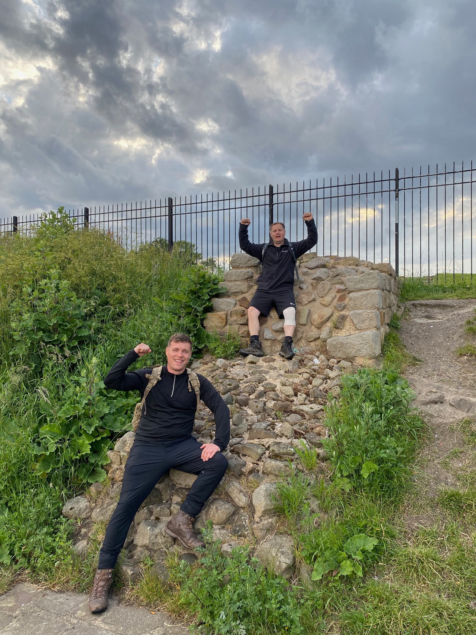 Two people pose outdoors near a rocky area with a metal fence behind them and a cloudy sky overhead. One is sitting on a stone structure with arms raised, while the other is crouching below, smiling and doing a similar pose.