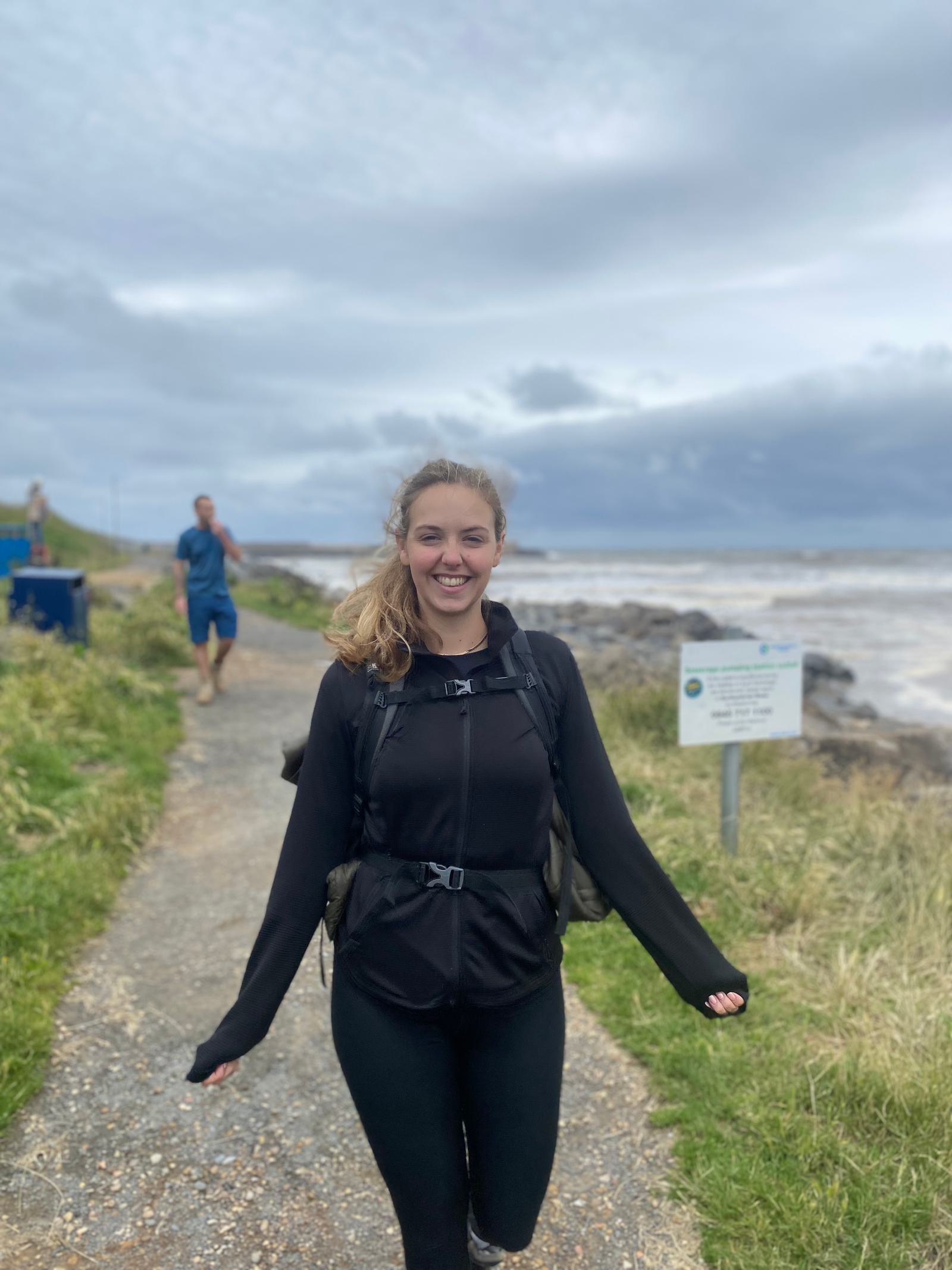A person in a black outfit walks on a coastal path, smiling. The sky is overcast, and the ocean is visible in the background. Another person walks behind on the path. A sign stands nearby amidst grassy surroundings.