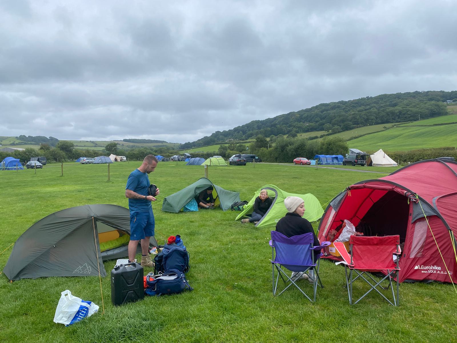 A group of people camping in a grassy field with scattered tents and camping gear. The scene includes rolling hills and a cloudy sky in the background. Some people sit in foldable chairs while others prepare beside the tents.