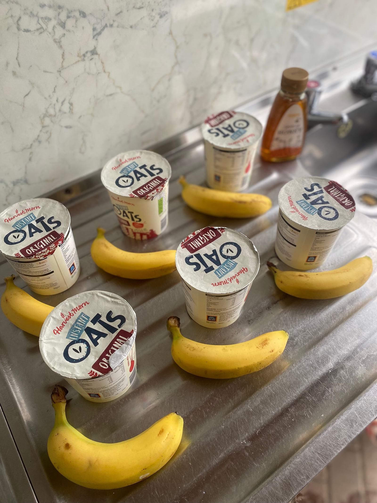 Five containers of overnight oats and five bananas are arranged on a stainless steel kitchen counter. A bottle of honey is visible in the background.