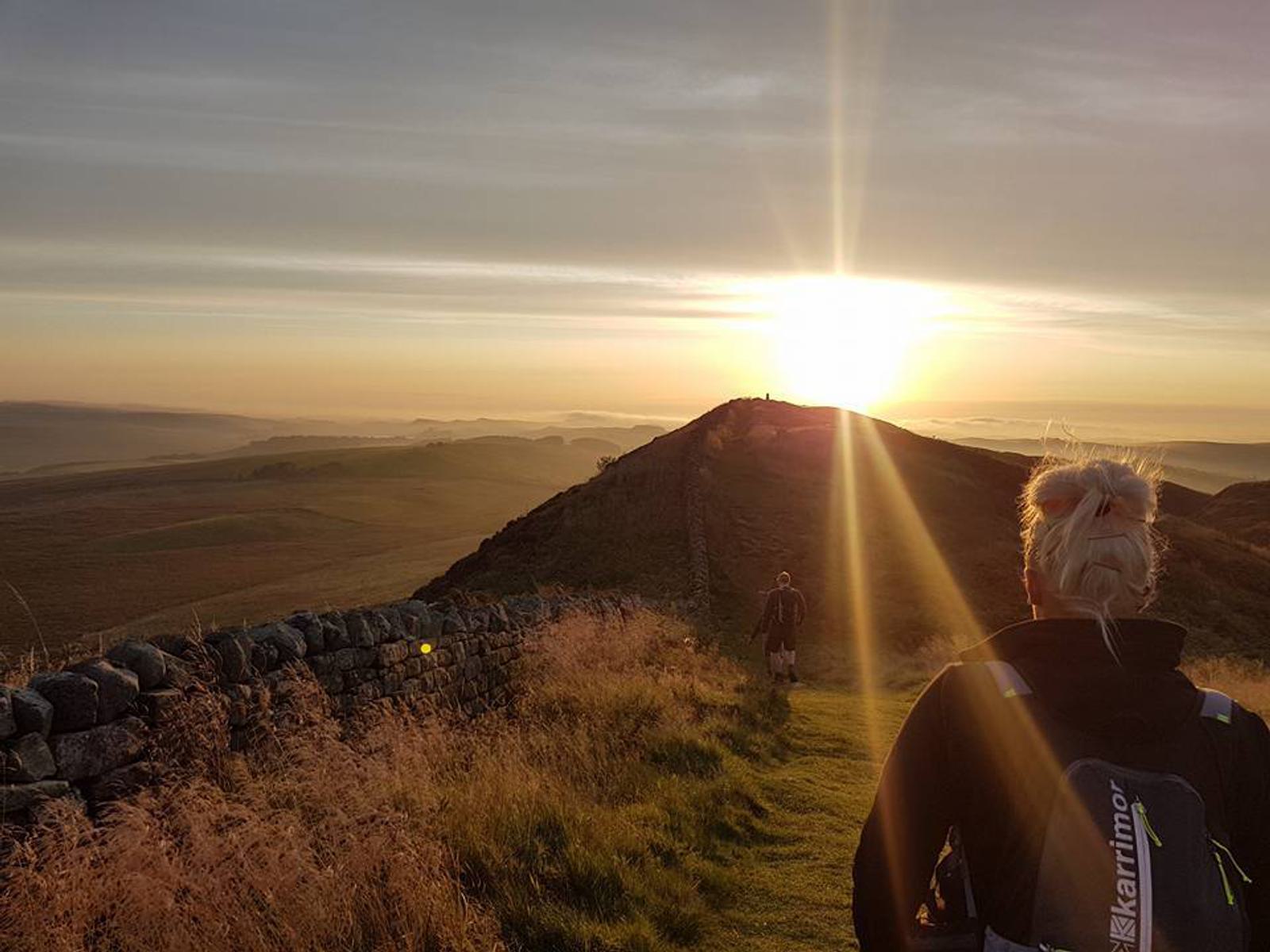 Two hikers walk along a grassy path at sunrise, with a stone wall to the left. The sun casts a warm glow over the hills and fields in the distance, creating a serene and picturesque landscape.