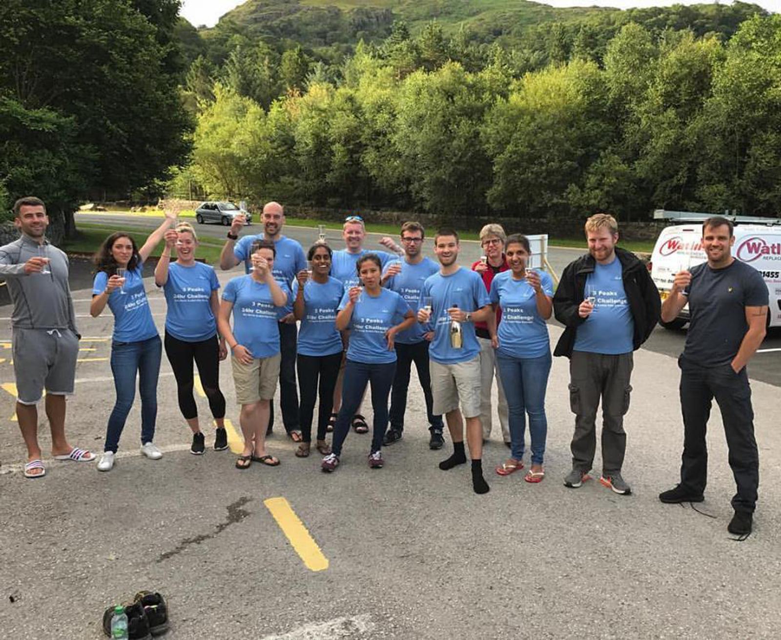 A group of fourteen people wearing matching blue T-shirts stands in a parking lot, smiling and holding drinks. Trees and a hill are visible in the background. A van with a logo is parked nearby.