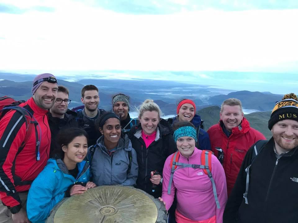 A group of ten people wearing outdoor clothing stand together smiling on a mountain summit. They are gathered around a metal marker under a cloudy sky, with distant hills and valleys visible in the background.
