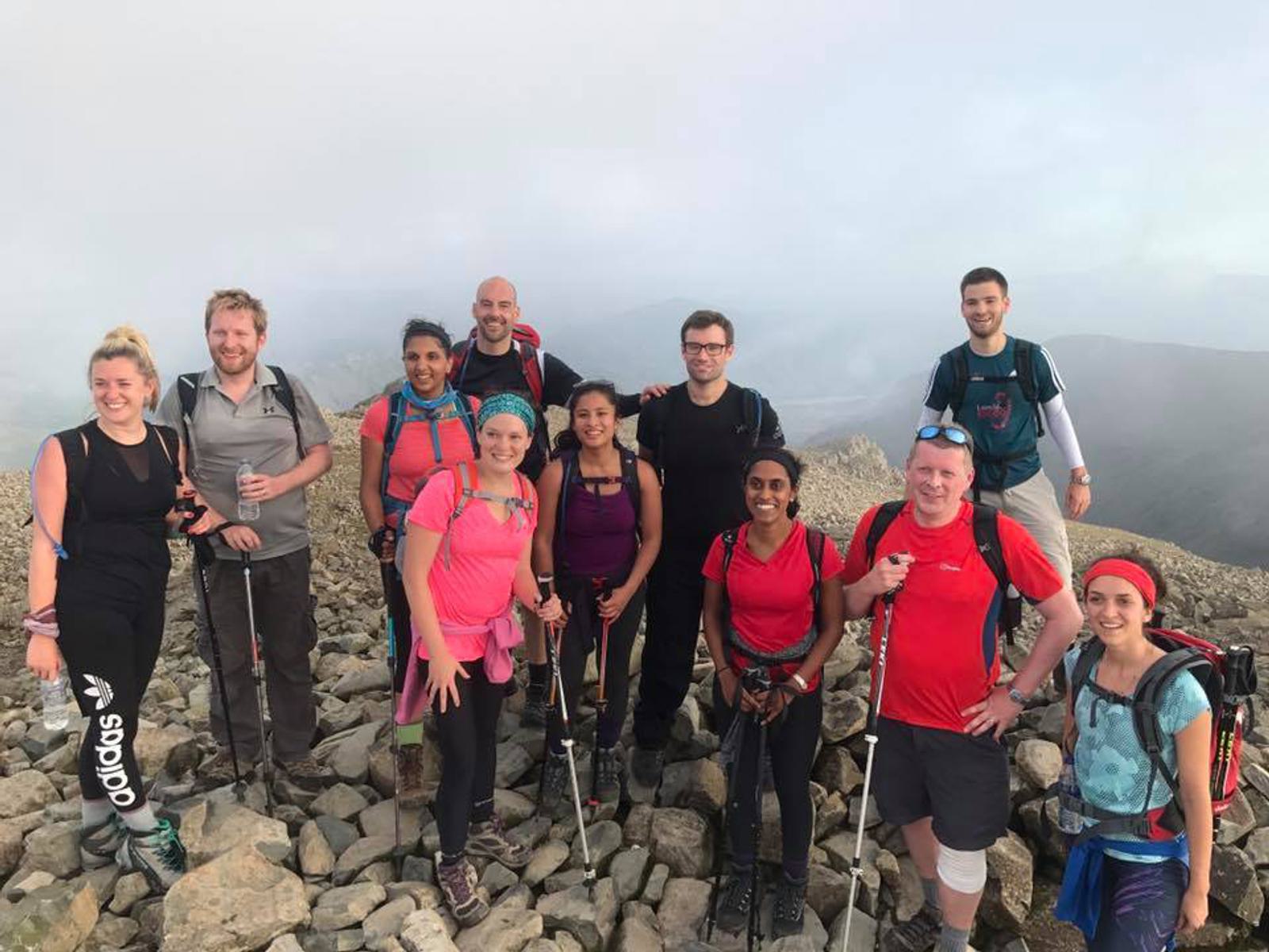 A group of ten people smiling and posing on a rocky mountain top during a hike. They are dressed in outdoor gear, with some holding hiking poles. The sky is cloudy, and mountains are visible in the background.