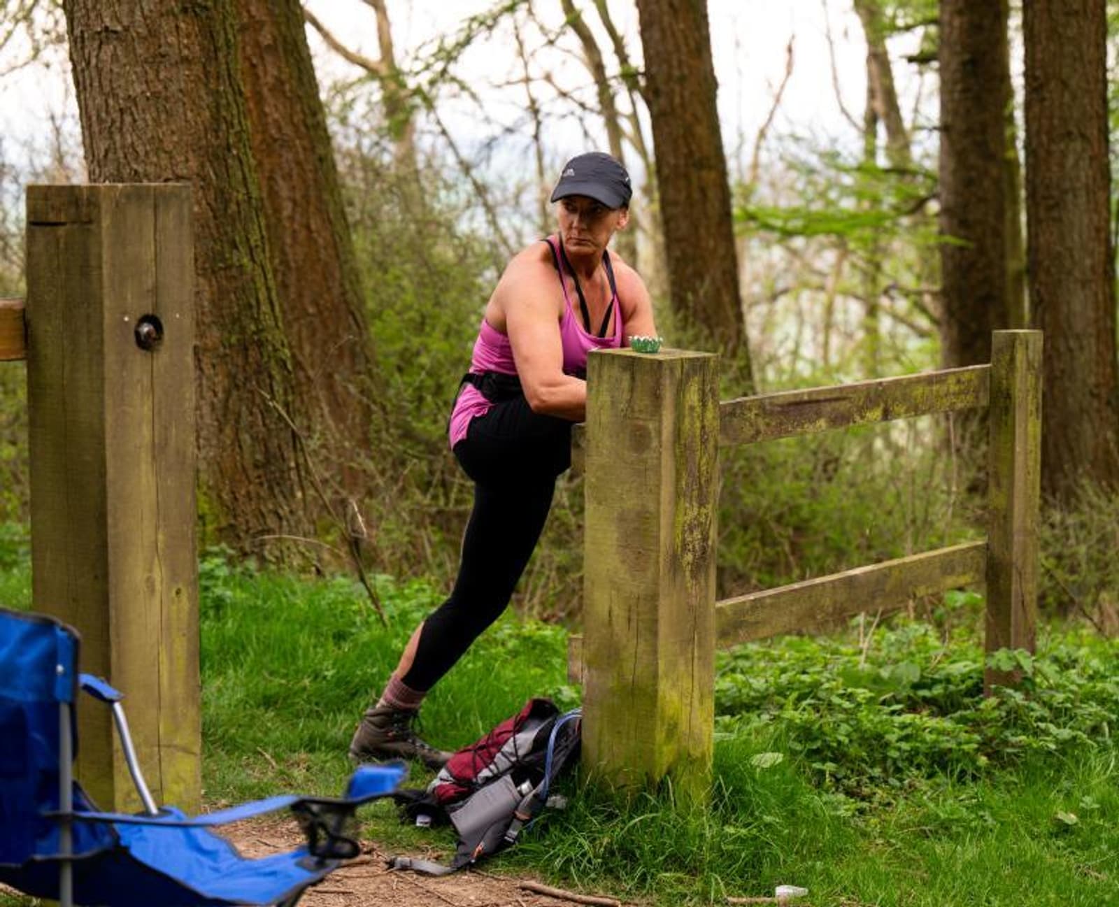 A woman in athletic clothing stretches her leg on a wooden fence in a forested area, with a backpack and folding chair nearby on the grassy ground.