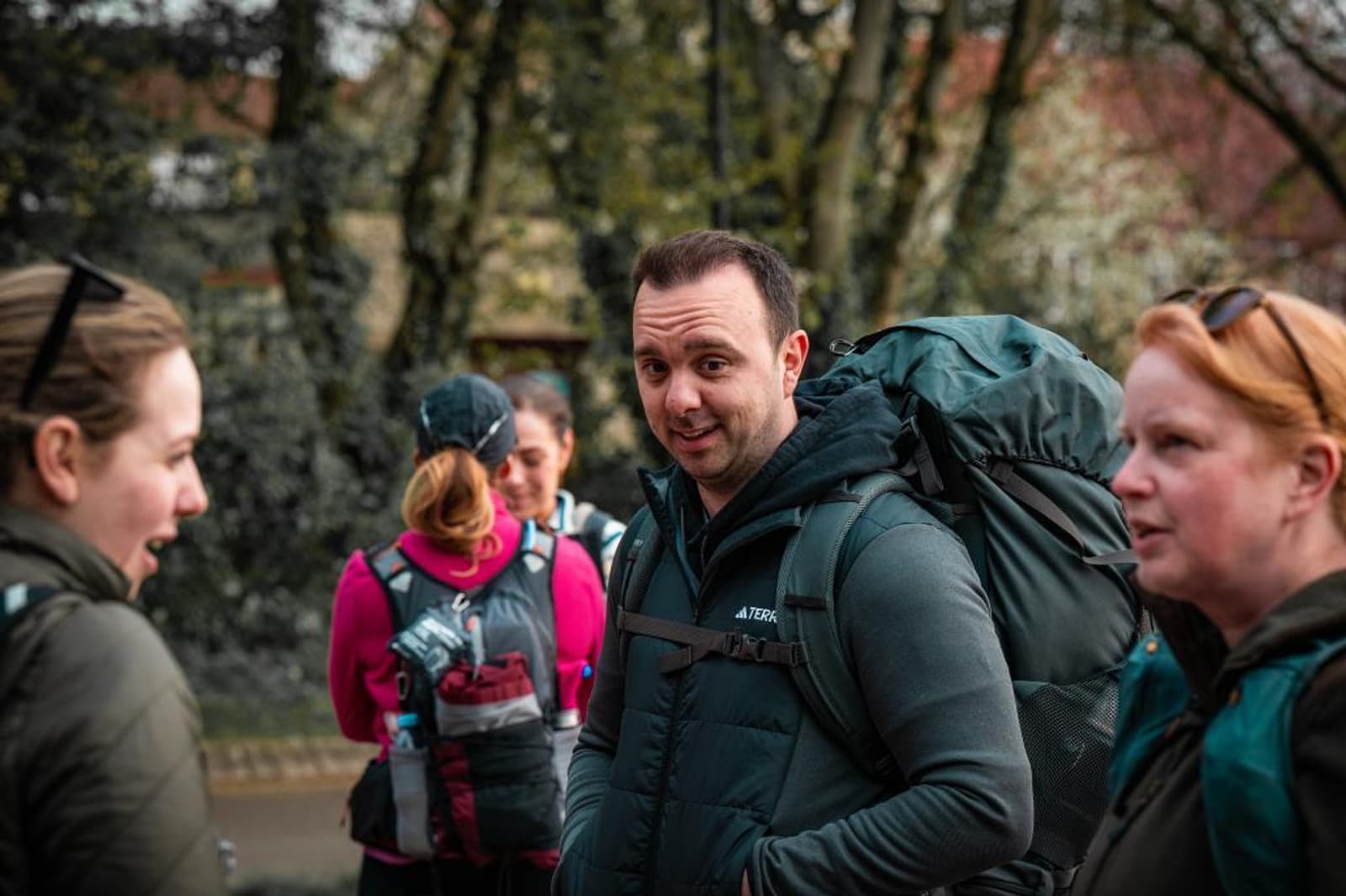 A man with a large backpack smiles at the camera while standing outdoors with three women, all dressed in outdoor gear. Trees and other greenery are visible in the background.