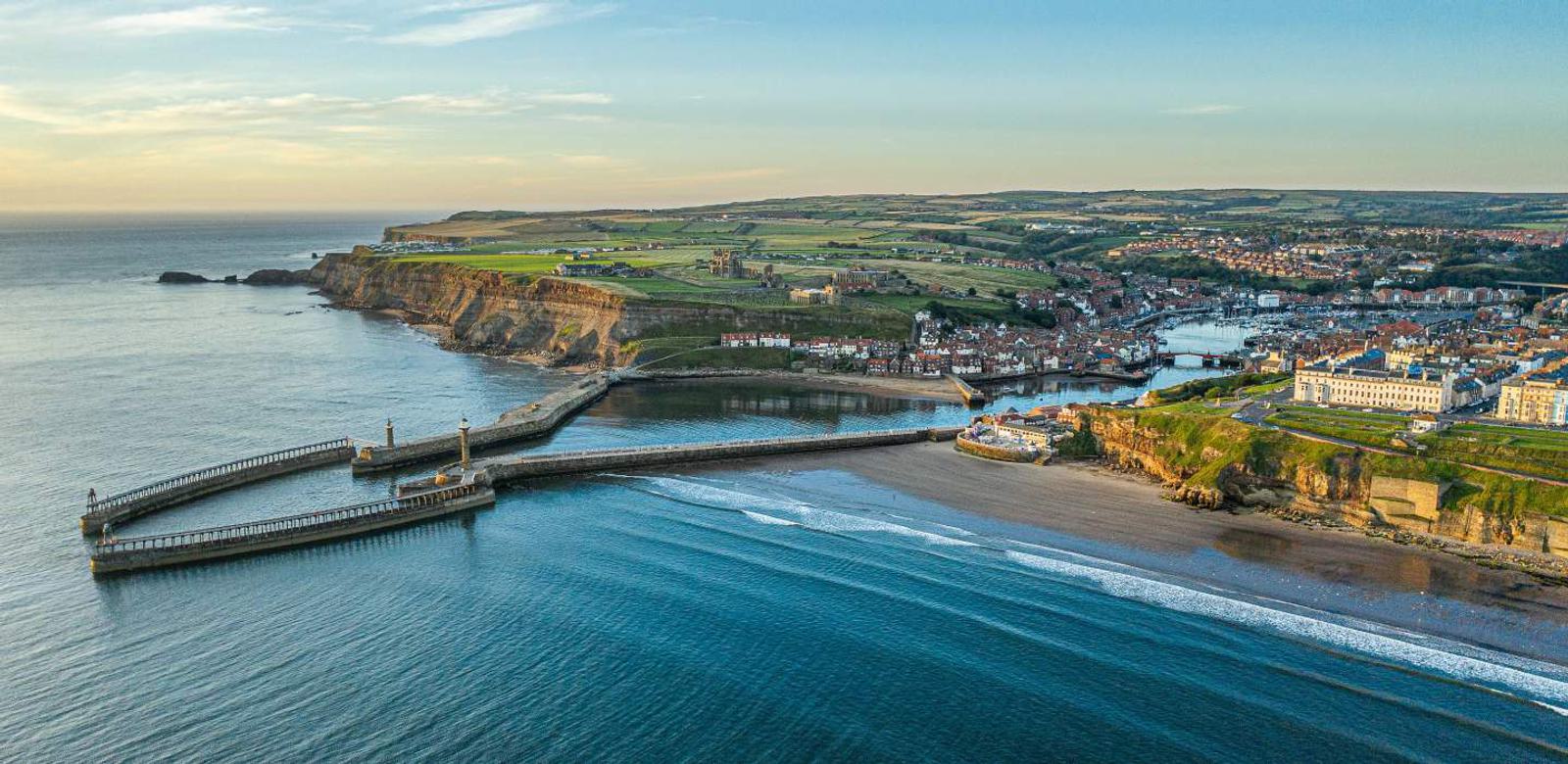 Aerial view of a coastal town with a large bay and long piers extending into the sea. The town is surrounded by green fields and cliffs. The ocean is calm, and the sky is partly cloudy with soft evening light.