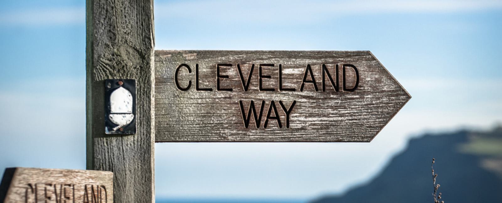 A wooden signpost with the text "Cleveland Way" points to the right. The post is weathered, adding a rustic feel. In the background, there is a blurred view of a coastal landscape under a clear blue sky.