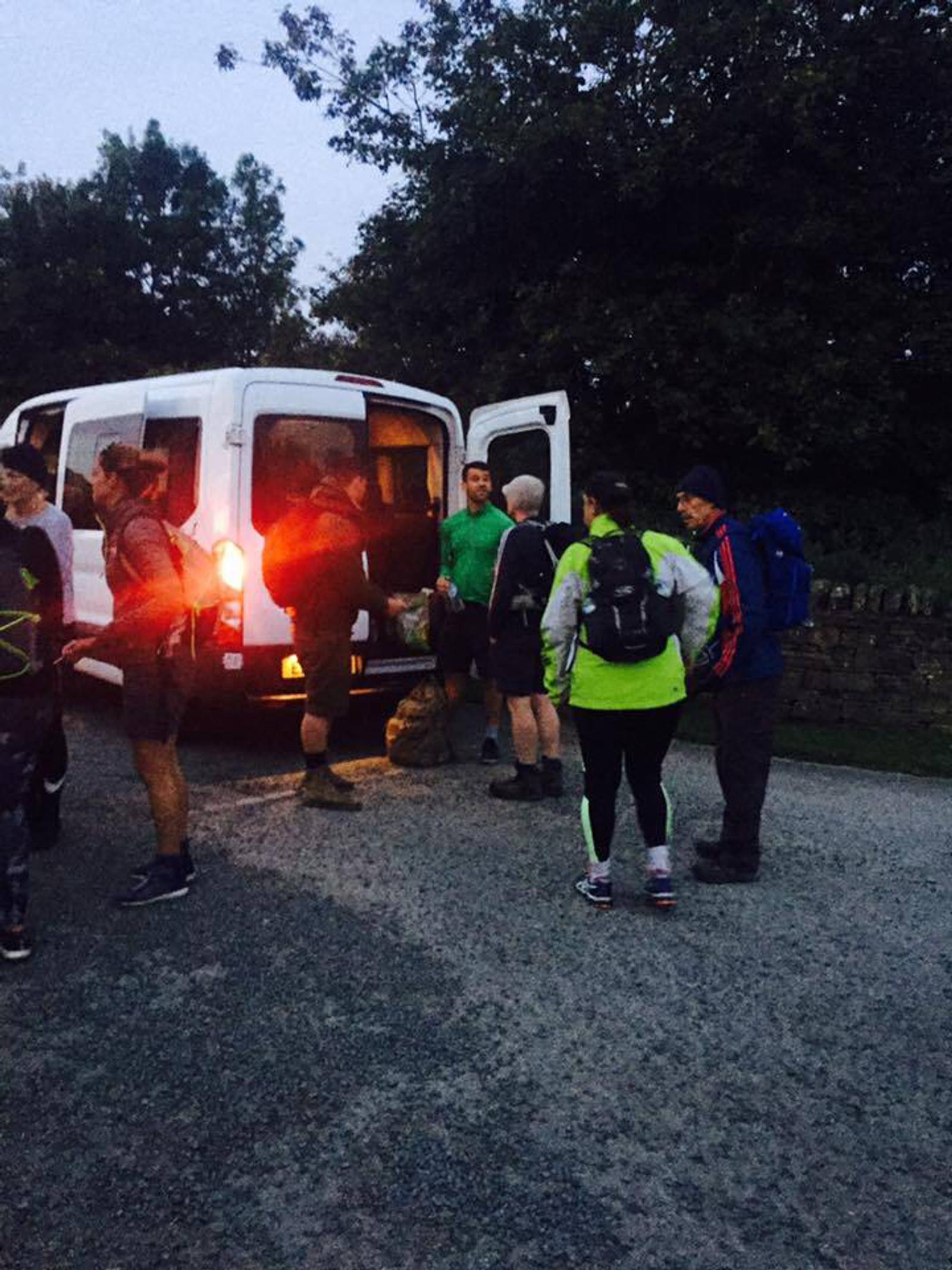 A group of hikers stands near a white van in a dimly lit parking area. Some are wearing backpacks and outdoor gear. The van's rear doors are open, and a light glows from inside. Trees and a low stone wall are visible in the background.