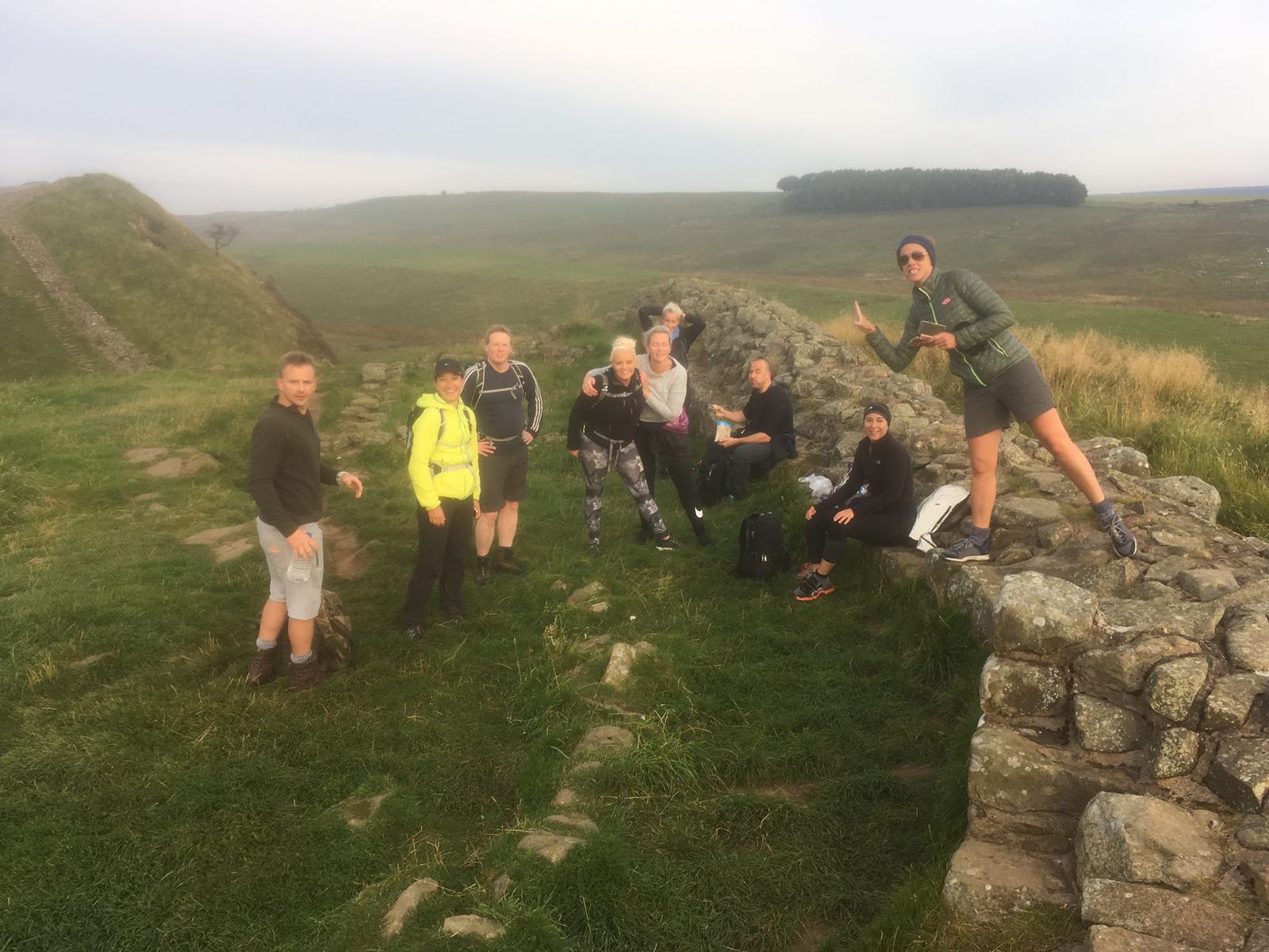 A group of hikers stands and sits on a stone path in a grassy area with one person playfully posing. They appear relaxed and happy, with rolling hills and a cloudy sky in the background.