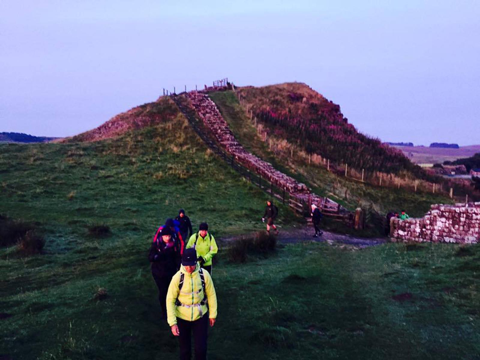 A group of hikers wearing bright jackets walks along a grassy path beside an ancient stone wall. The sky is overcast, and a small hill in the background is partially covered with grass and a stone staircase.