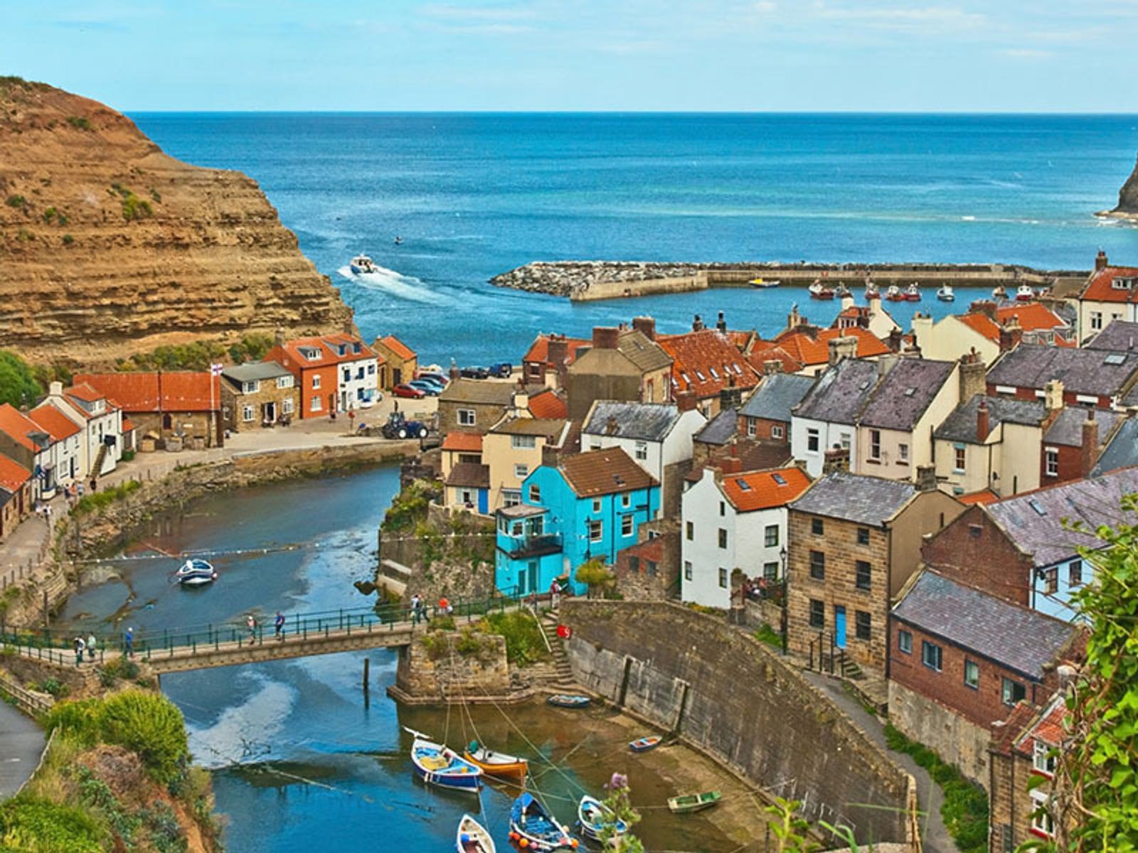A picturesque coastal village with a collection of colorful houses nestled by the sea. Boats are moored along the narrow waterway, surrounded by steep cliffs. The open sea stretches into the horizon under a cloudy sky.