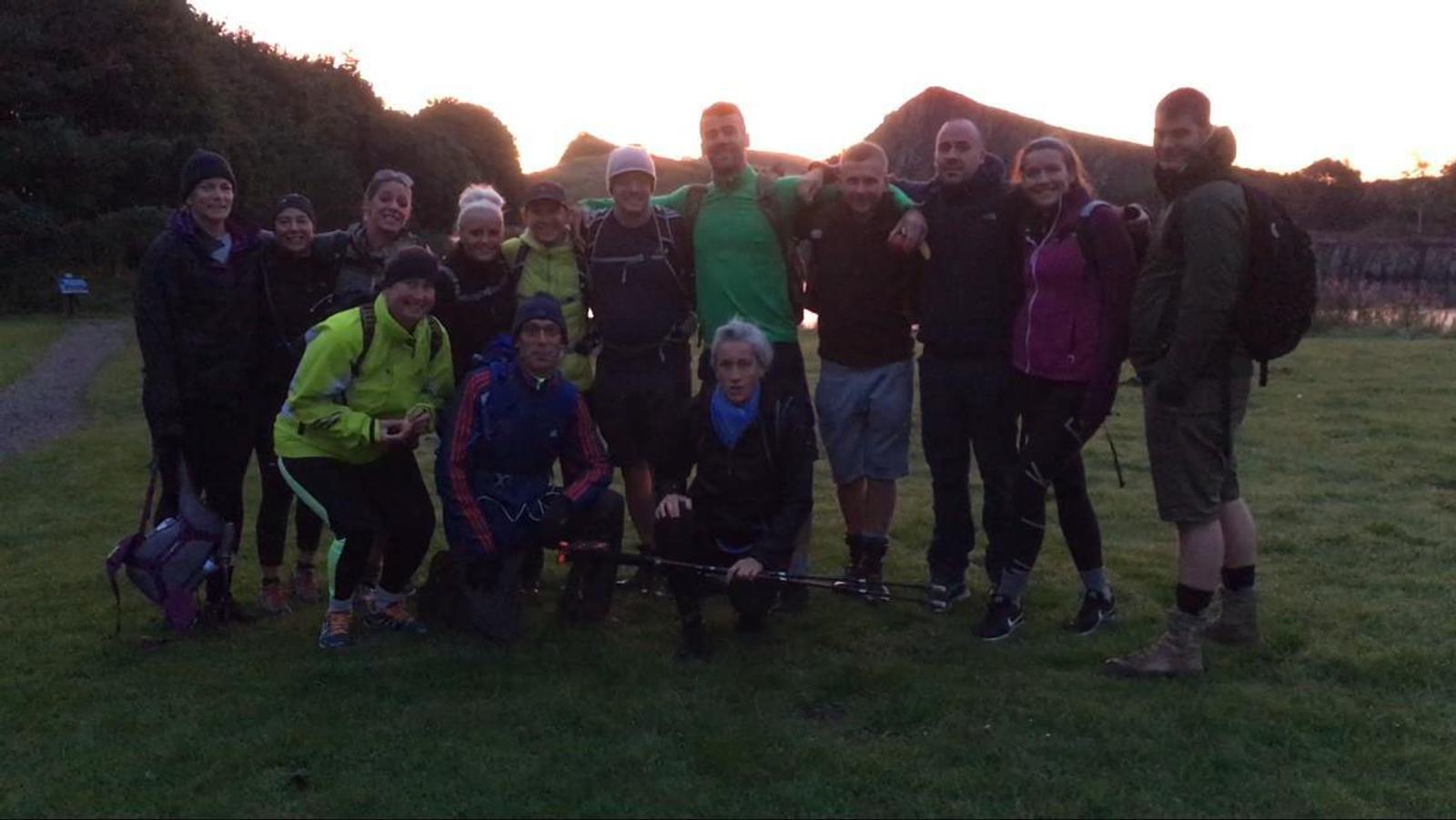 A group of people dressed in outdoor gear pose together on a grassy area at dawn or dusk. They are smiling and appear to be ready for a hike, with a dimly lit sky and hill in the background. Some hold hiking poles and backpacks are visible.
