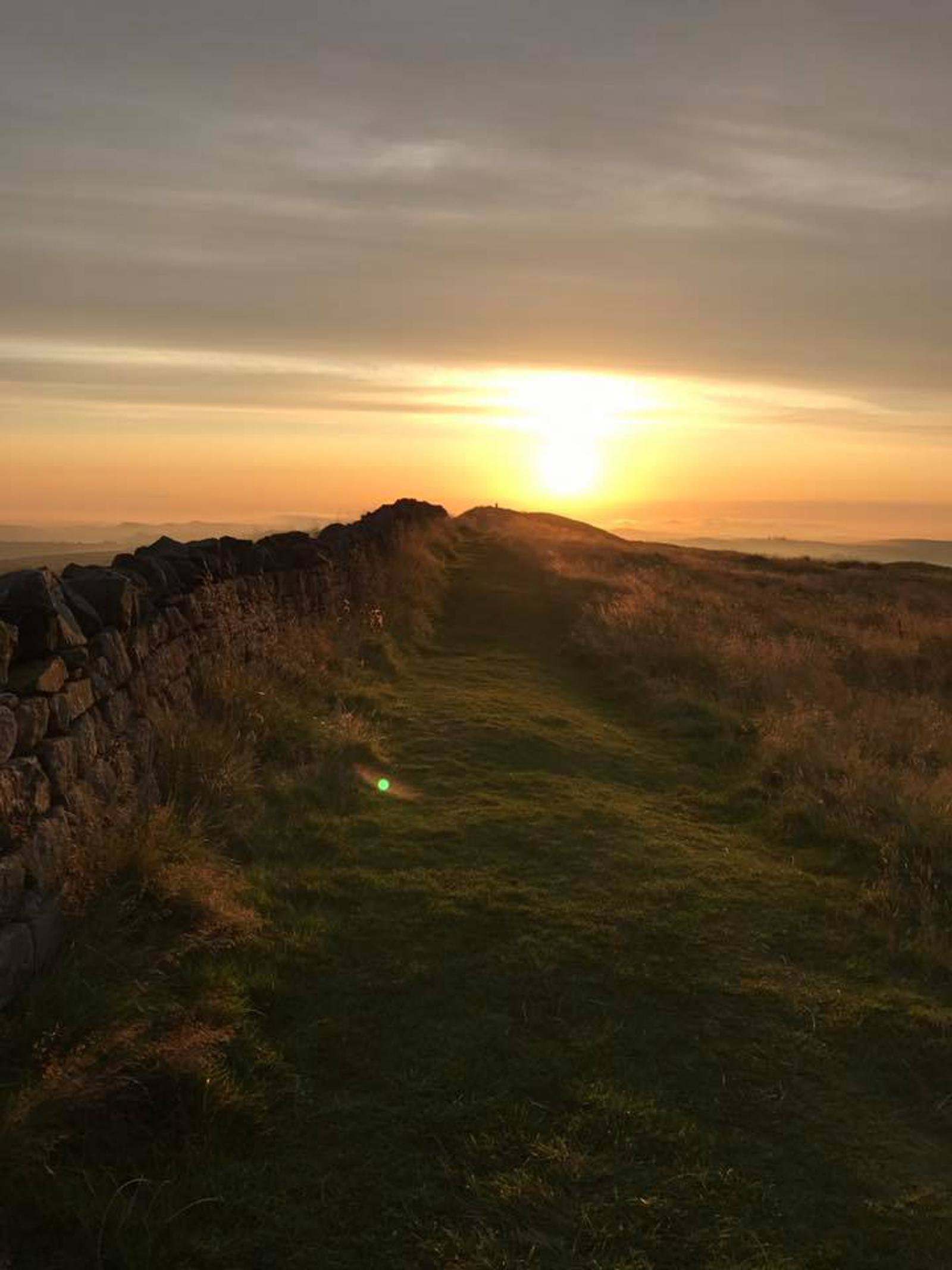 A serene landscape with a stone wall lining a grassy path under a golden sunrise. The sky is filled with soft clouds, creating a tranquil early morning atmosphere over the rolling hills.