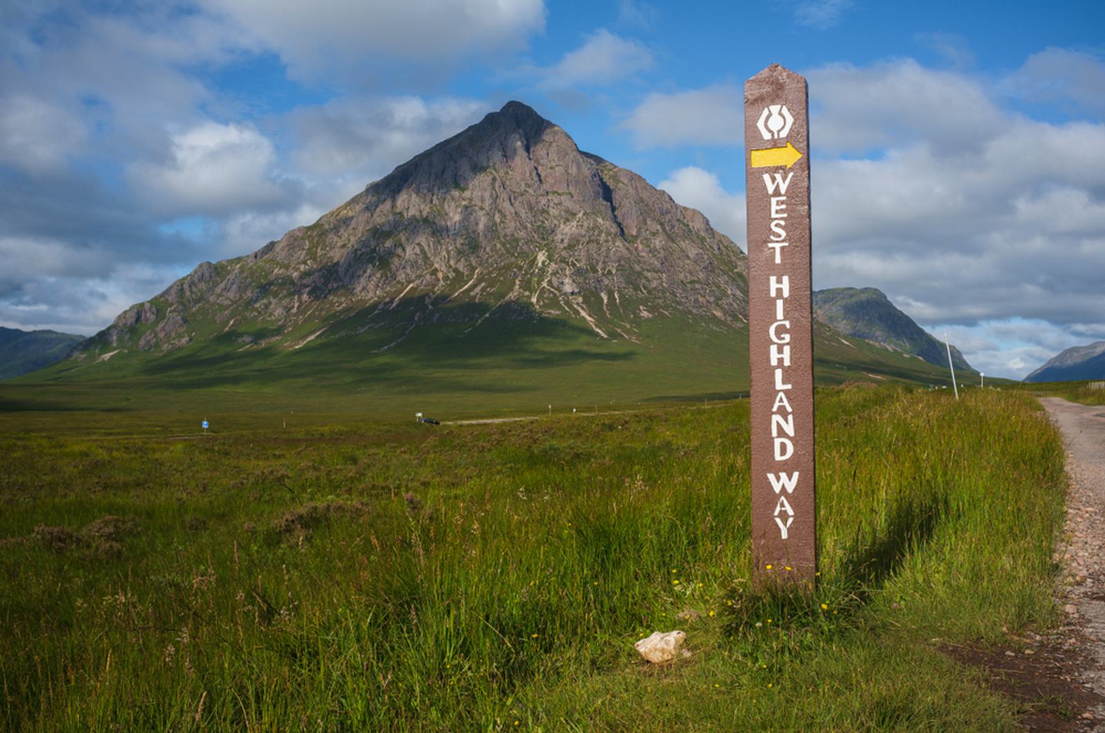 A wooden signpost reading "West Highland Way" stands in a grassy field under a blue sky. In the background, a large, rocky mountain rises, partially covered in shadow.