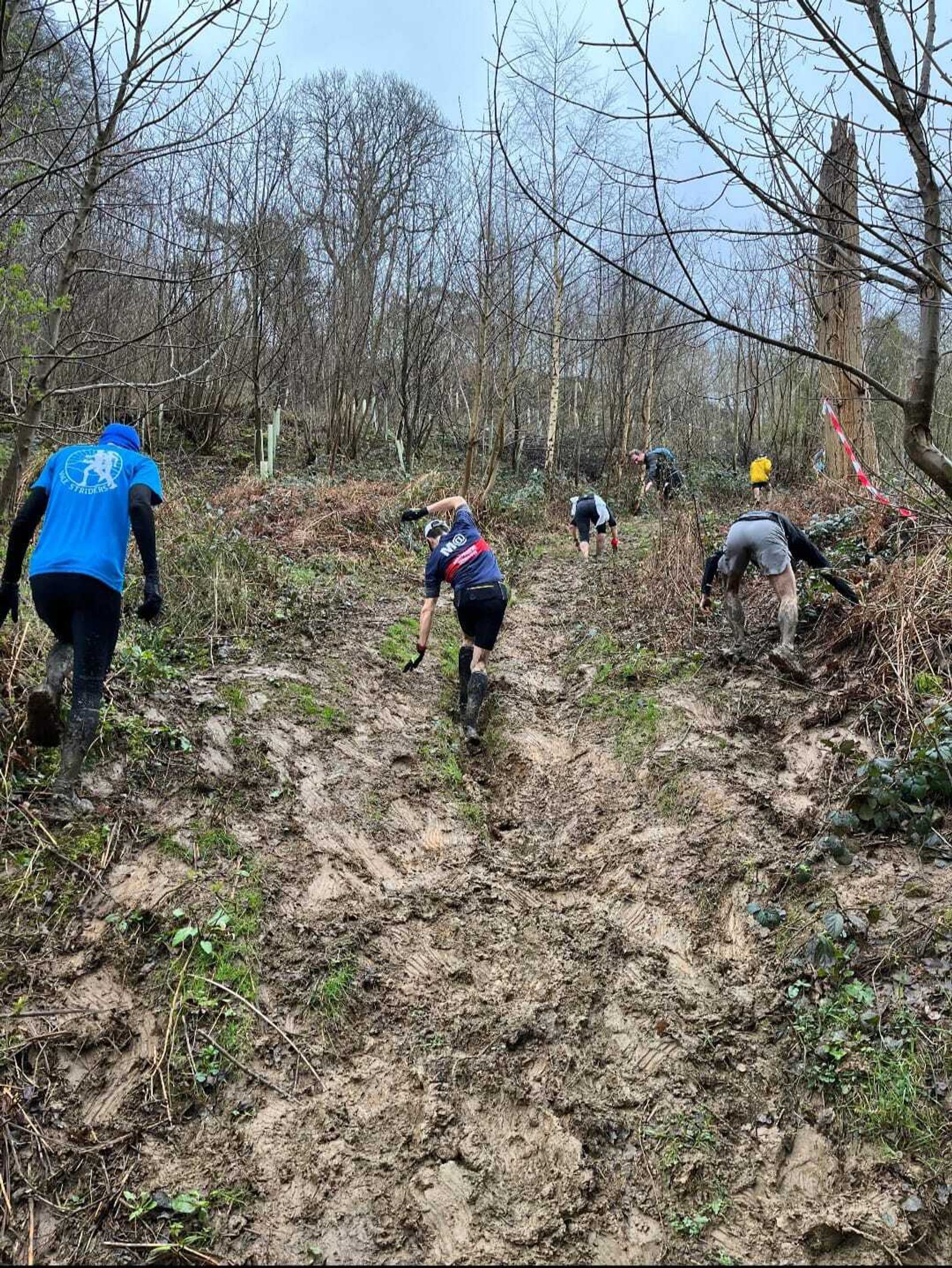 Participants climb a steep, muddy hill in a forest during an outdoor race. They are wearing athletic clothing and navigating through a challenging, slippery terrain. Trees surround the area, and the sky is overcast.