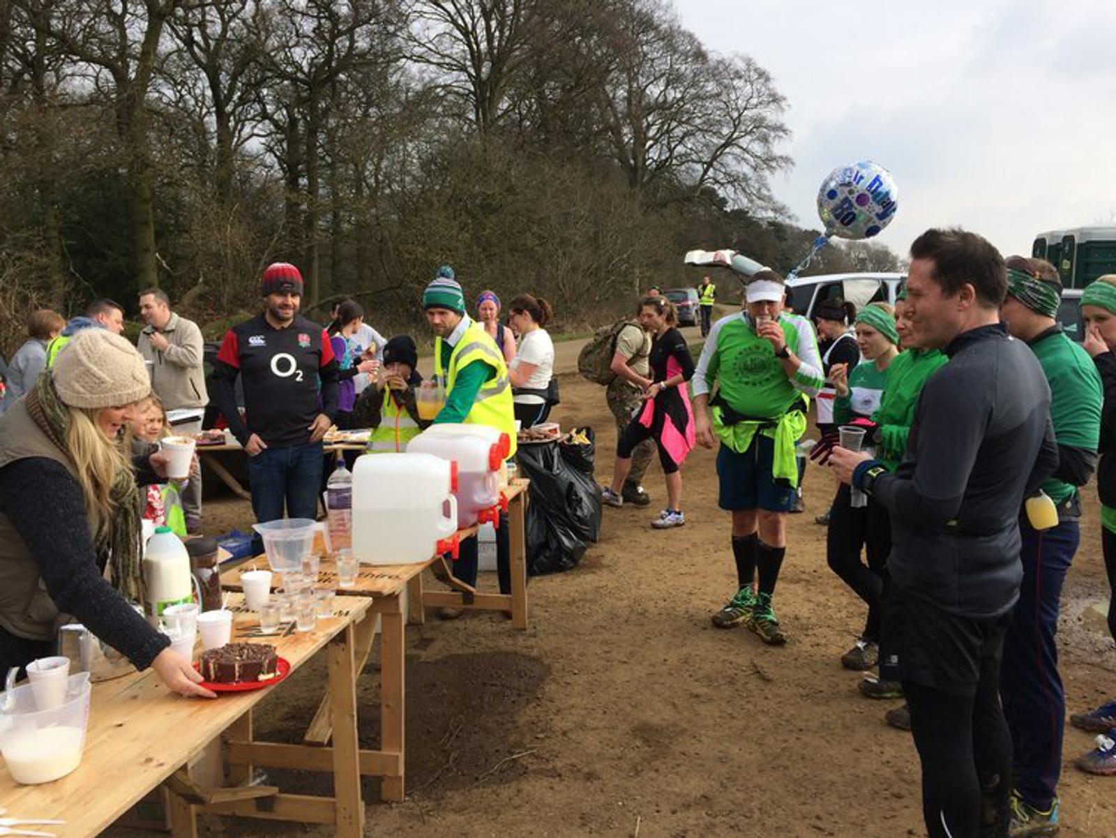 A group of runners gather at a refreshment table in a park during an outdoor event. Volunteers serve drinks and snacks. Some participants wear green outfits, and one person holds a balloon. Trees and a vehicle are visible in the background.