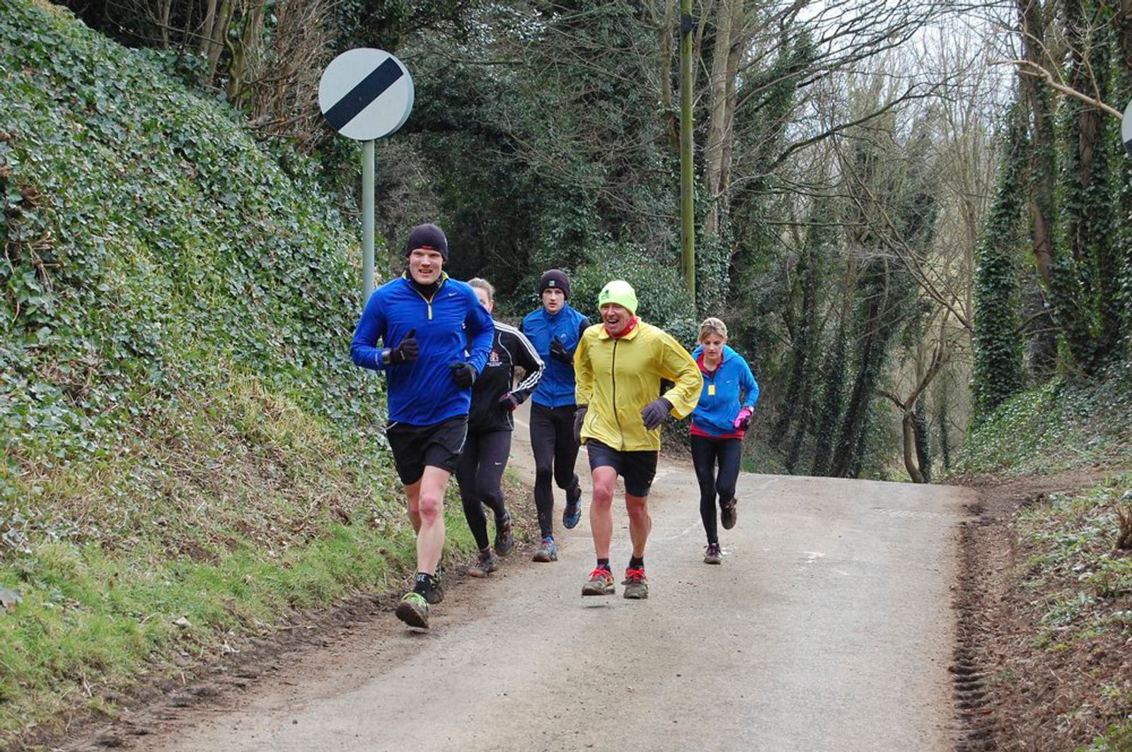 A group of five runners jogs uphill on a narrow road surrounded by greenery. They wear winter gear, including jackets and hats. A circular road sign is visible on the left. The scene suggests a chilly, outdoor workout.