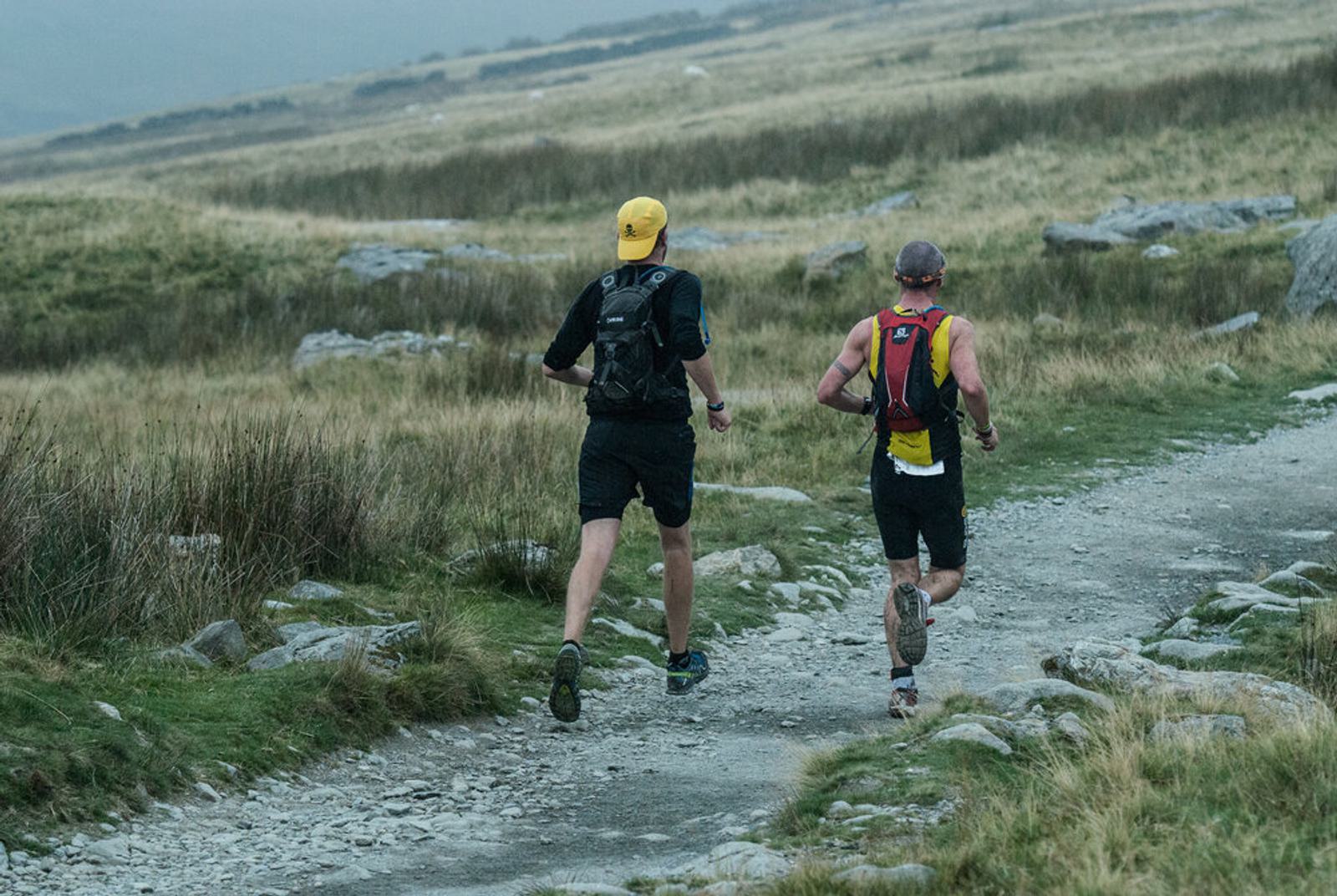 Two men trail running on a rocky path through a grassy landscape. Both wear hats and backpacks; one has a yellow cap and black shorts, the other a burgundy cap and yellow singlet. The sky is overcast, creating a moody atmosphere.