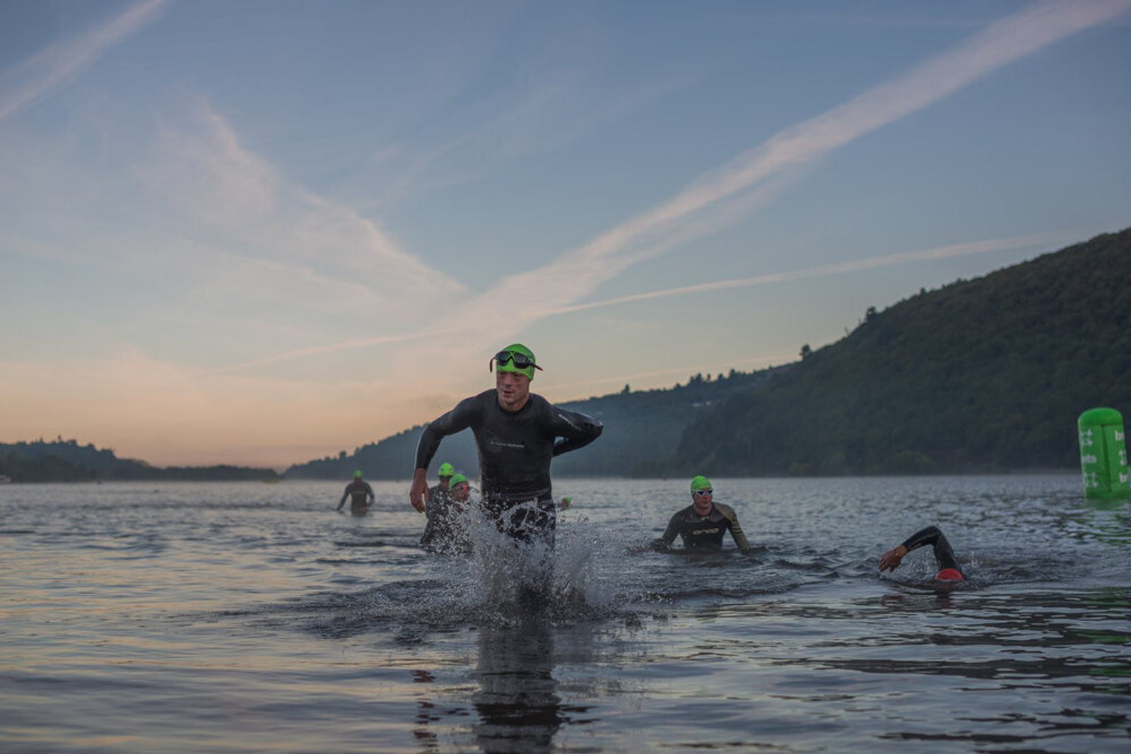 A group of triathletes in wetsuits and green swim caps emerge from the water during an early morning race. The lead athlete is running through the shallow water, with others following behind against a backdrop of hills and a clear sky.