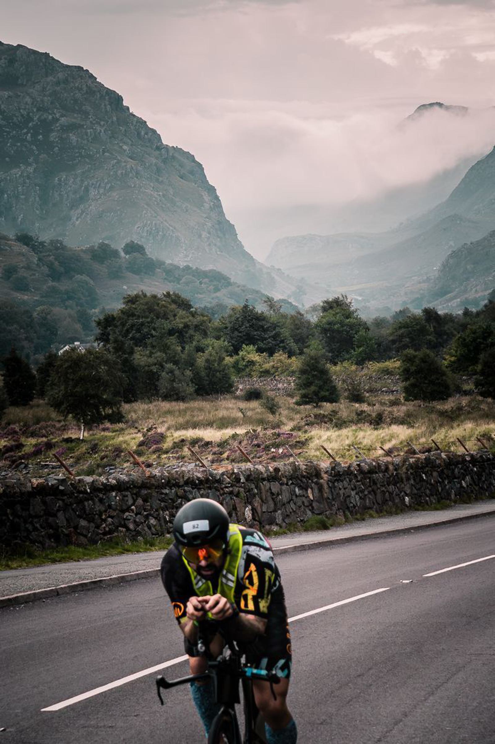 A cyclist in racing gear rides a bike on a road surrounded by lush greenery and rolling hills. Majestic mountains in the background are partially shrouded in mist under a cloudy sky, creating a dramatic and serene landscape.