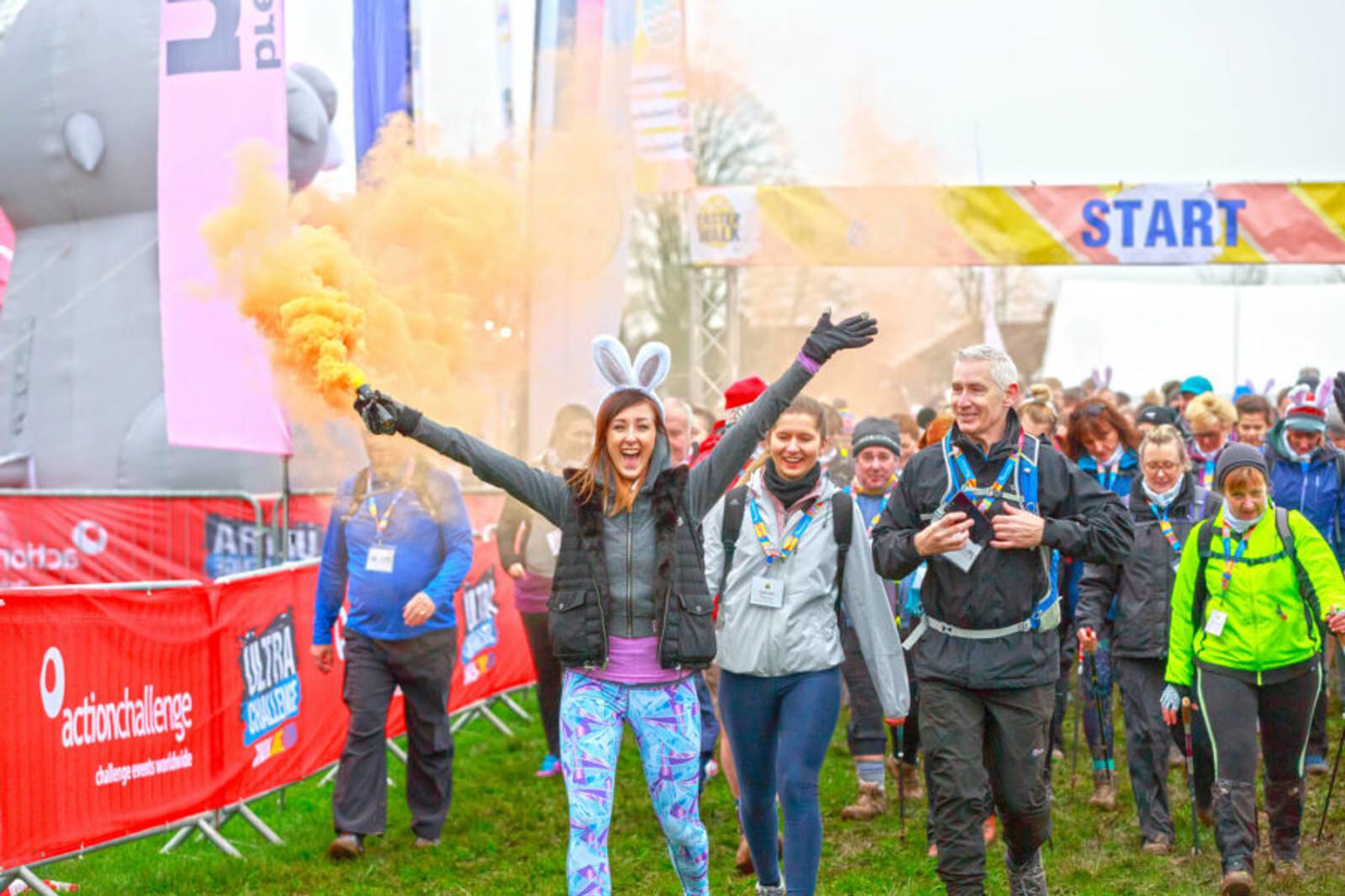 A group of people participate in an outdoor charity walk. The lead woman, wearing bunny ears, joyfully holds an orange smoke flare. Others follow behind, some smiling and wearing hiking gear. A large inflatable bear and start banner are visible in the background.