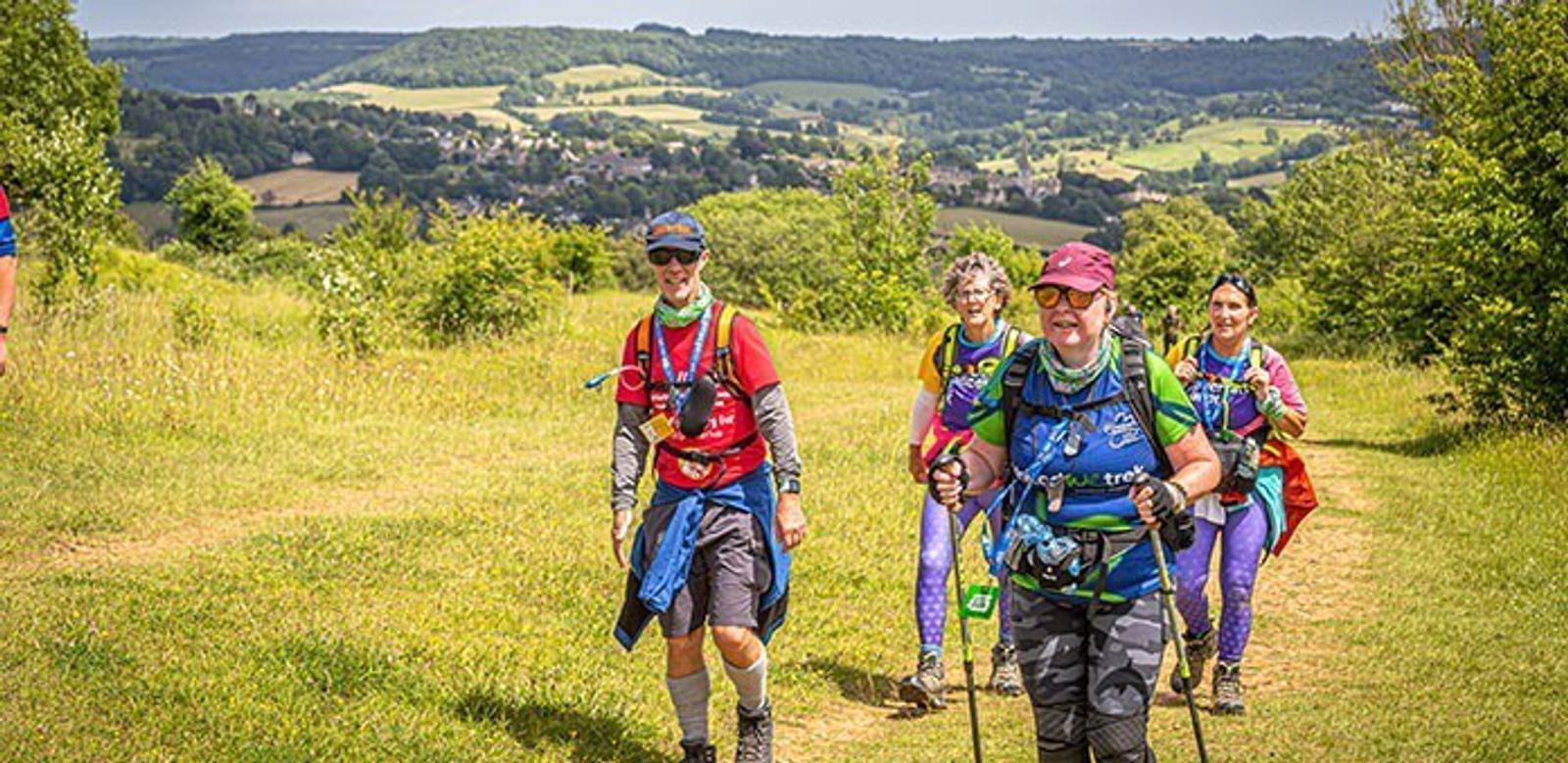 A group of four people hiking on a trail with lush green hills in the background. They are wearing colorful athletic outfits and carrying backpacks. The sky is clear, and the scene is bright and sunny.