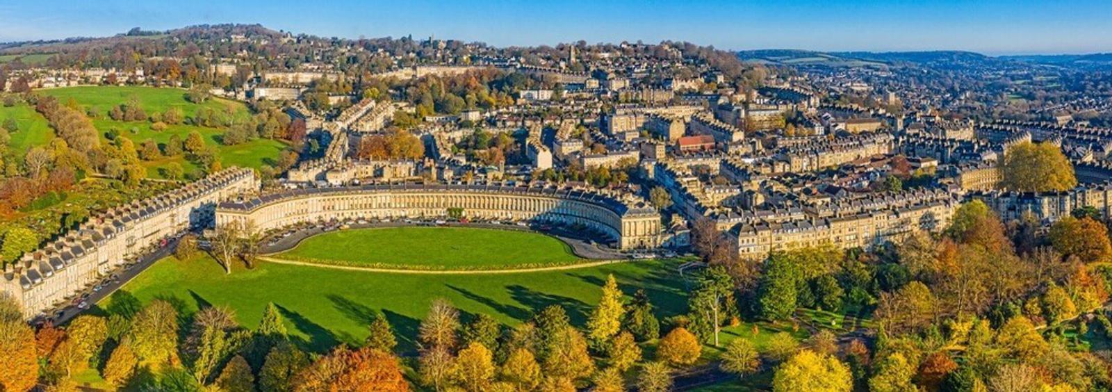 Aerial view of the Royal Crescent in Bath, UK, a sweeping row of Georgian terraced houses. Set amidst lush green fields and autumnal trees, the historic cityscape extends into the distance under a clear blue sky.