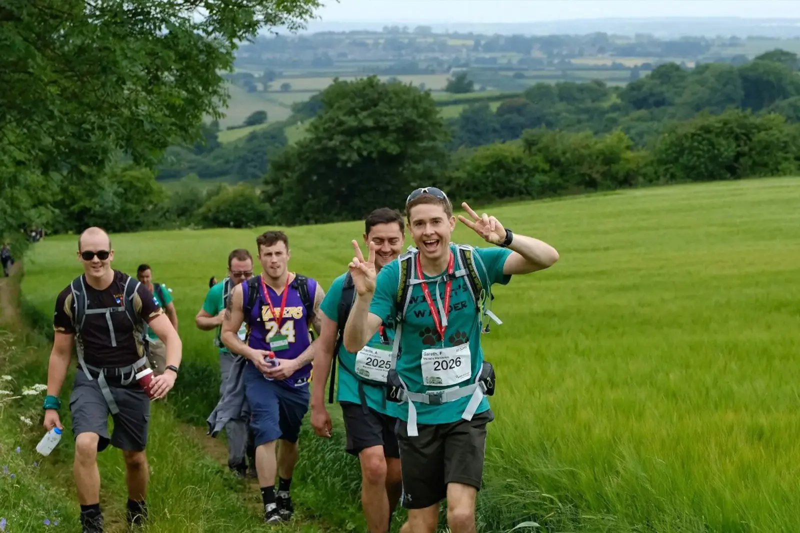 A group of six people hiking along a scenic trail with green fields and distant hills. A man in the foreground smiles and gestures with peace signs. They are carrying backpacks and wearing casual athletic clothing.