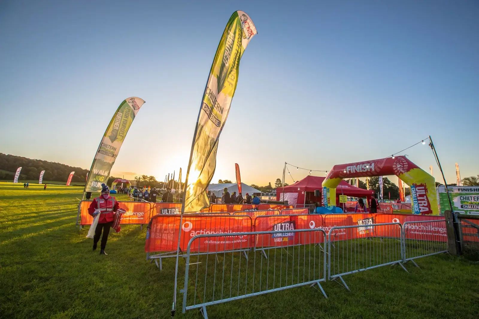 Sunrise over a grassy outdoor event area with tall banners and a red inflatable finish line arch. Fenced pathways guide participants. A person in a red jacket walks nearby. The scene conveys the start or end of a race in a vibrant setting.