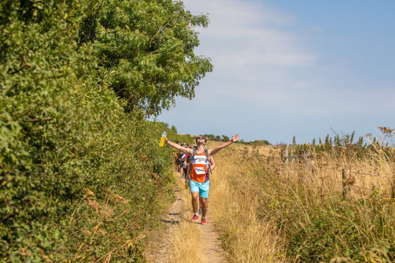 A runner in a race on a narrow dirt path between green shrubs and dry grass raises their arms triumphantly. They hold a yellow water bottle. The sky is clear and sunny, with others following in the background.