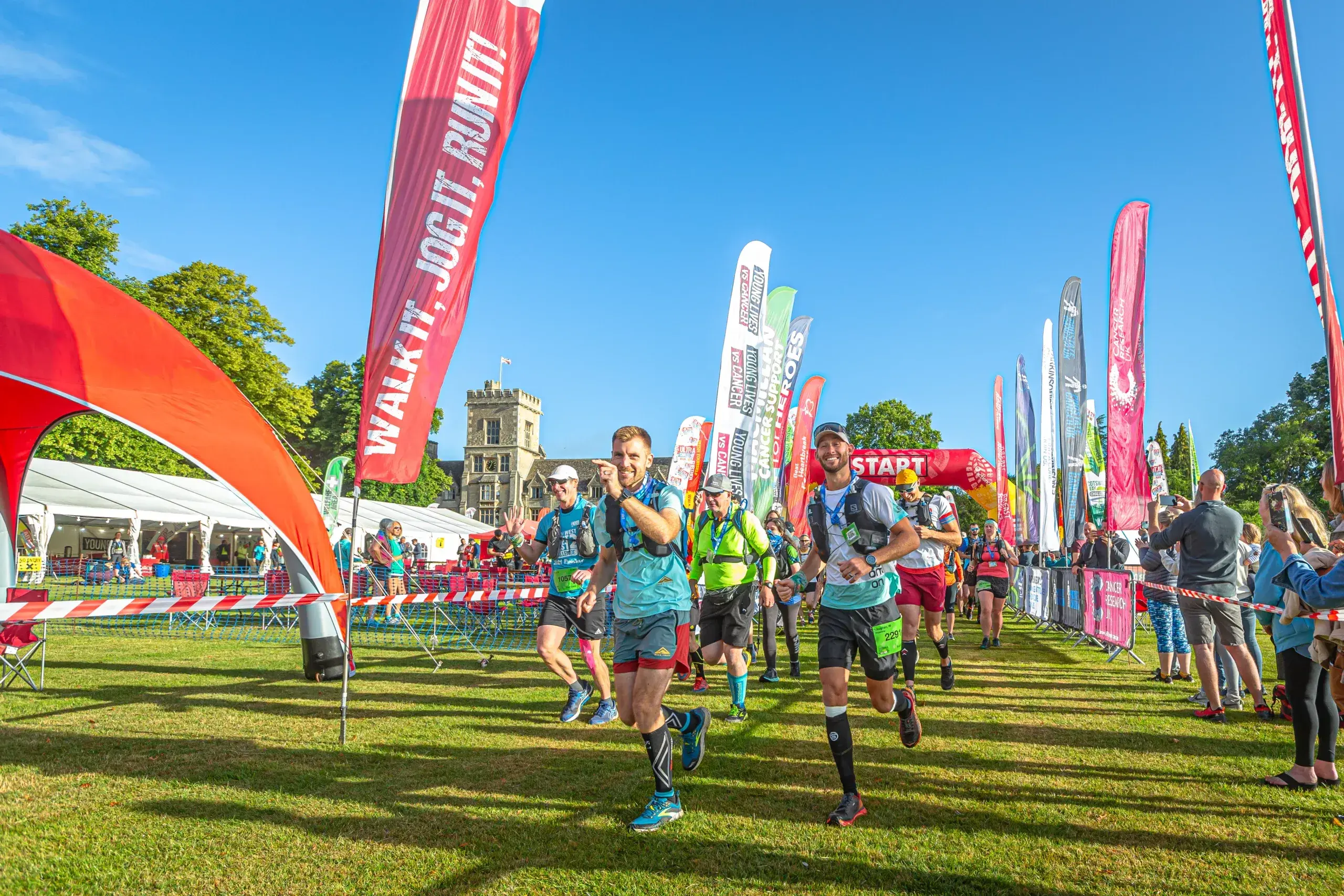 A group of runners at a race event, starting under a red arch with various colorful flags and banners in the background. The event takes place on a sunny day with a clear blue sky and a historic building visible. Spectators stand nearby.
