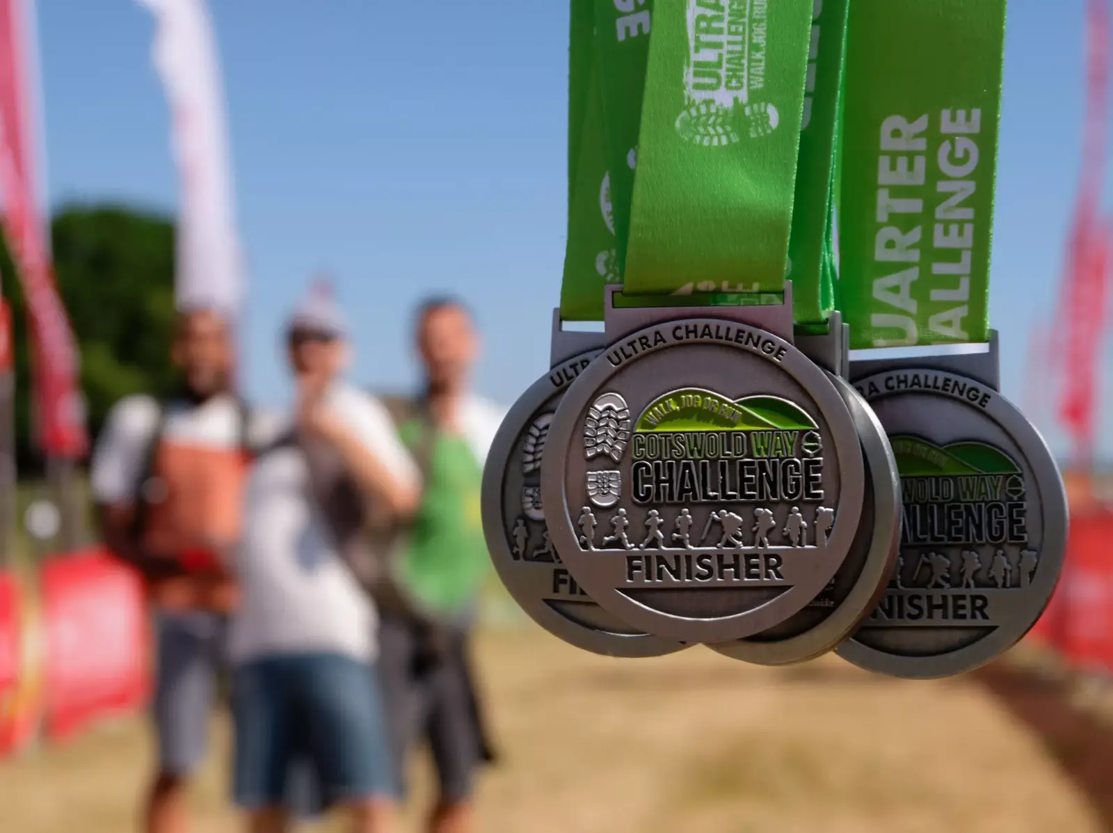 Close-up of three silver medals with green ribbons reading "Cotswold Way Challenge Finisher." The background shows three people standing on a grassy field with red banners, slightly out of focus.