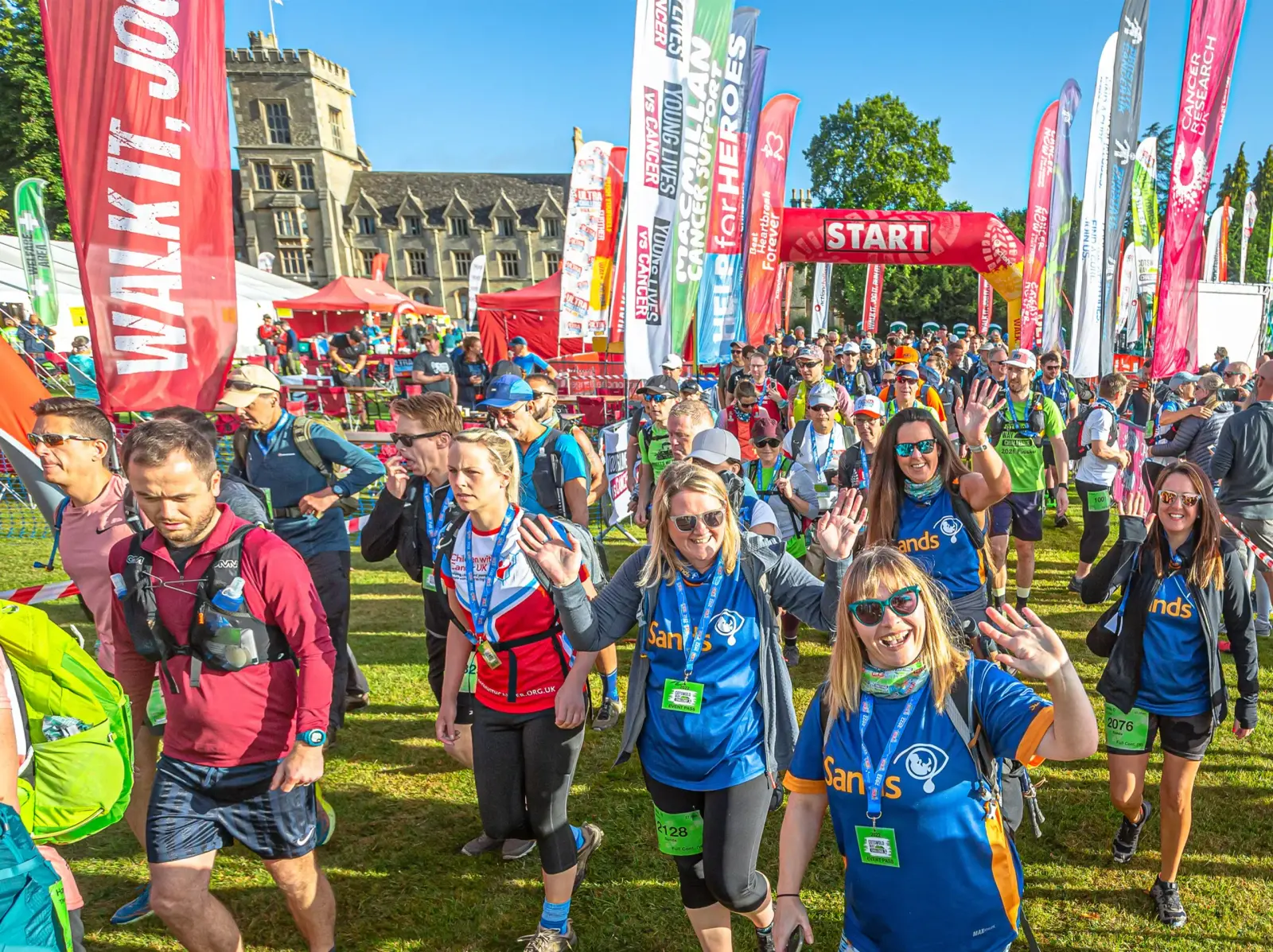 A large group of people participating in an outdoor event, walking past a starting line. They are wearing colorful shirts and walking gear. Banners and a red "START" sign are visible, along with a scenic building and trees in the background.