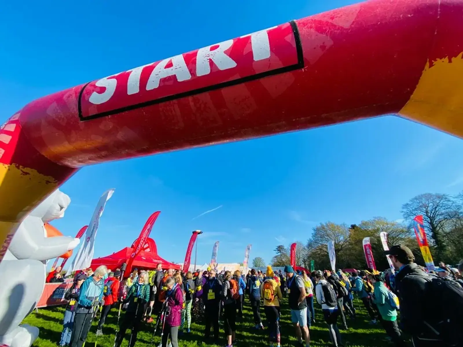 A crowd of participants gathers under a red and yellow inflatable arch marked "START" in a park. The sky is clear and blue, and there are colorful flags and a red tent in the background.